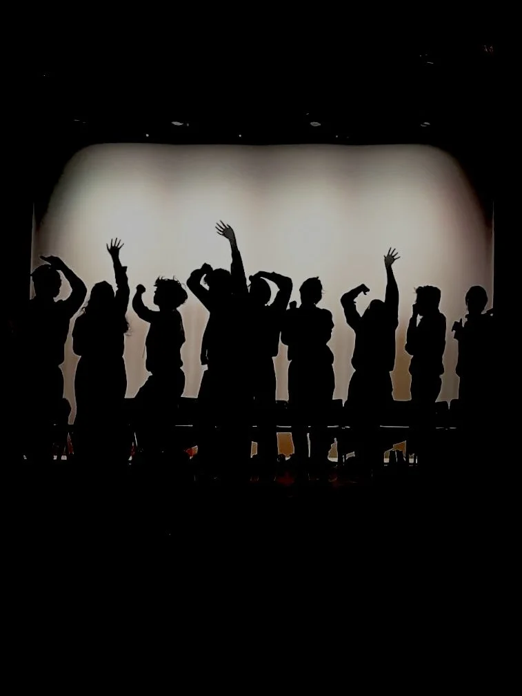 Silhouettes de plusieurs personnes debout sur scène, certaines levant les bras ou les mains dans des positions variées, avec un fond lumineux et sombre.