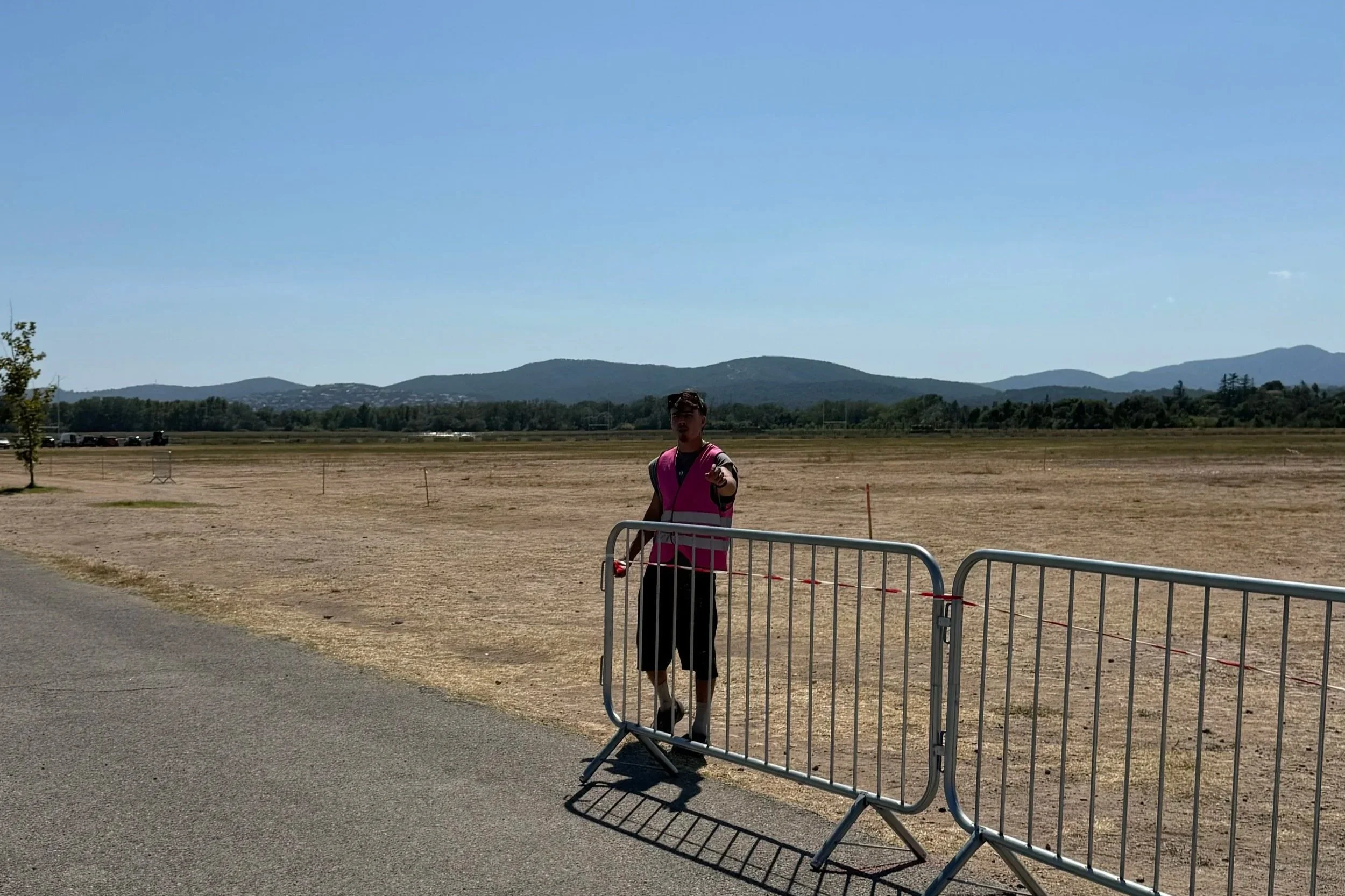 Jeune homme portant un gilet rose et une casquette, pointant du doigt devant un panneau de barrière dans un espace ouvert avec des montagnes en arrière-plan, sous un ciel clair.
