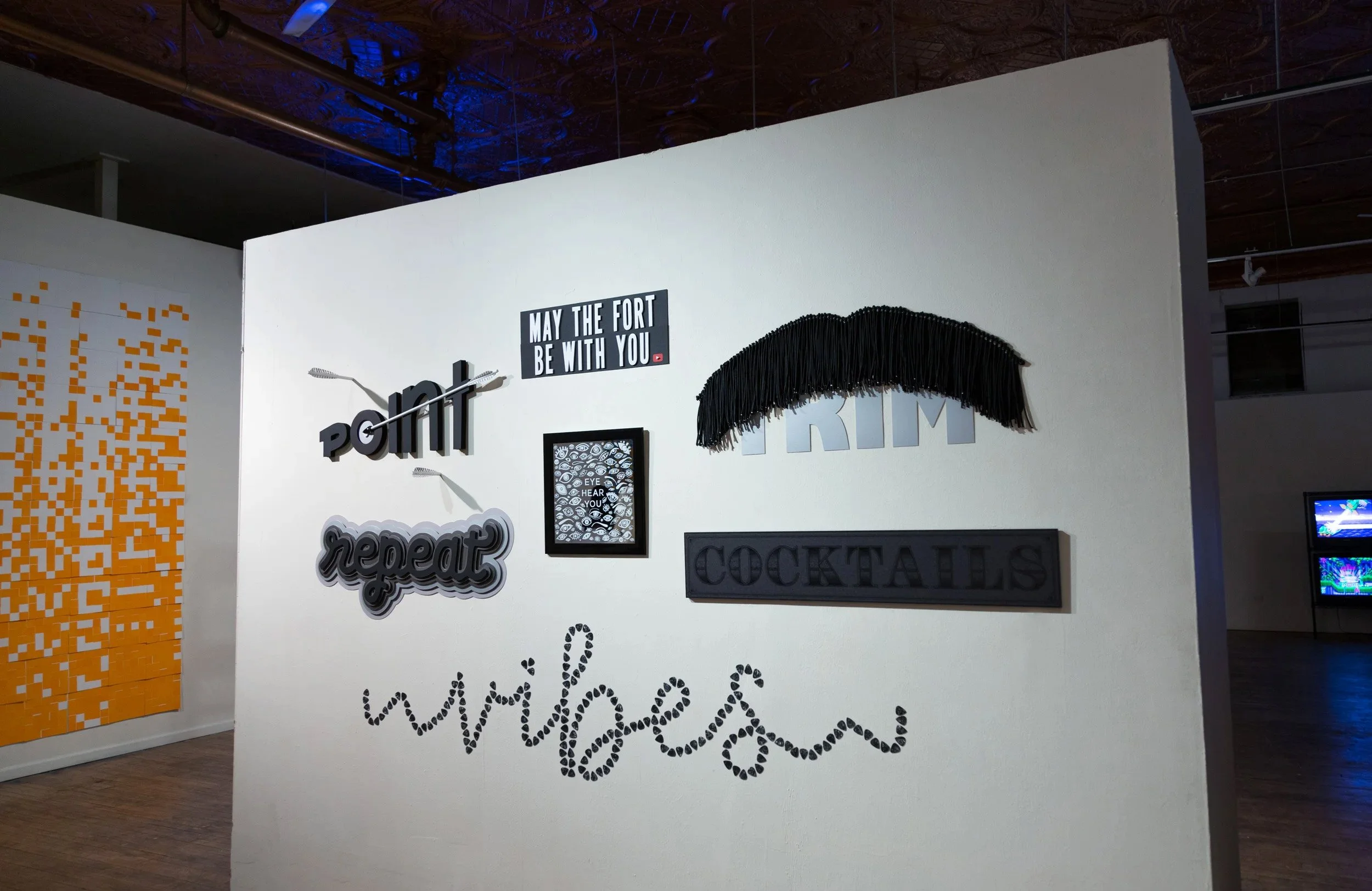 A white gallery wall displaying various art and decorative signs, including phrases like 'point', 'repeat', 'may the fort be with you', 'TRIM', 'COCKTAILS', and 'whimbeads' in different fonts and styles, with a black fringe decoration and a smaller framed eye design.