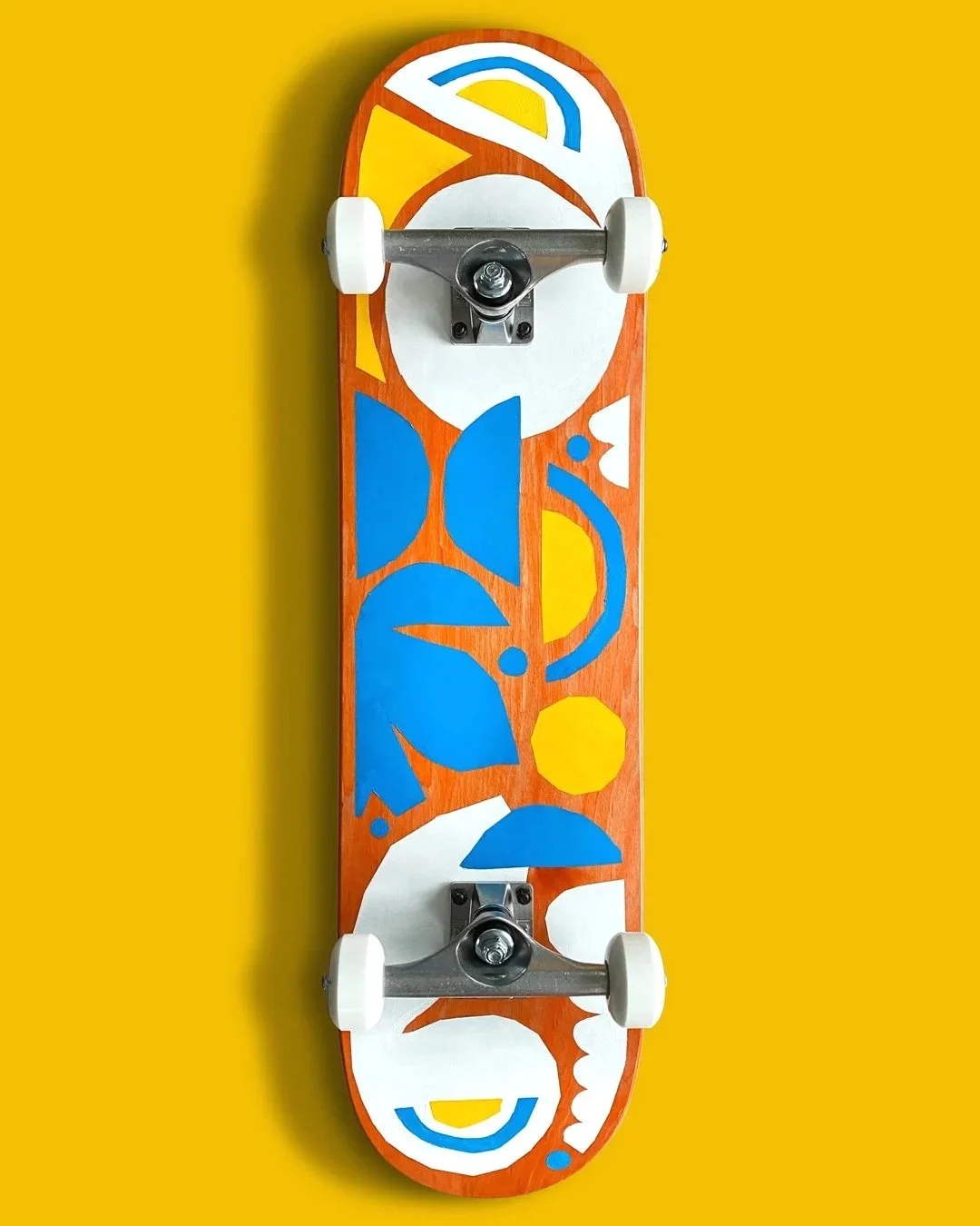 A skateboard with a colorful abstract design featuring blue, yellow, white, and orange shapes, mounted on a yellow wall.