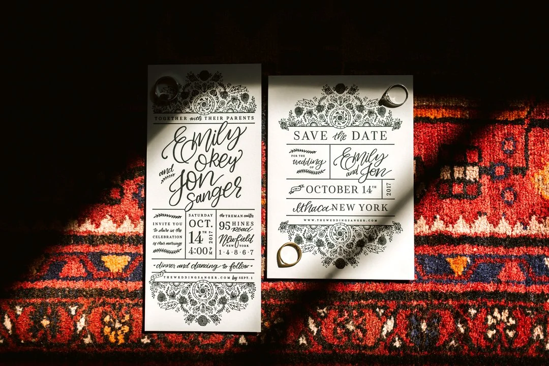 Two wedding save the date cards placed on a decorative red and blue patterned rug, with rings on top of the cards.