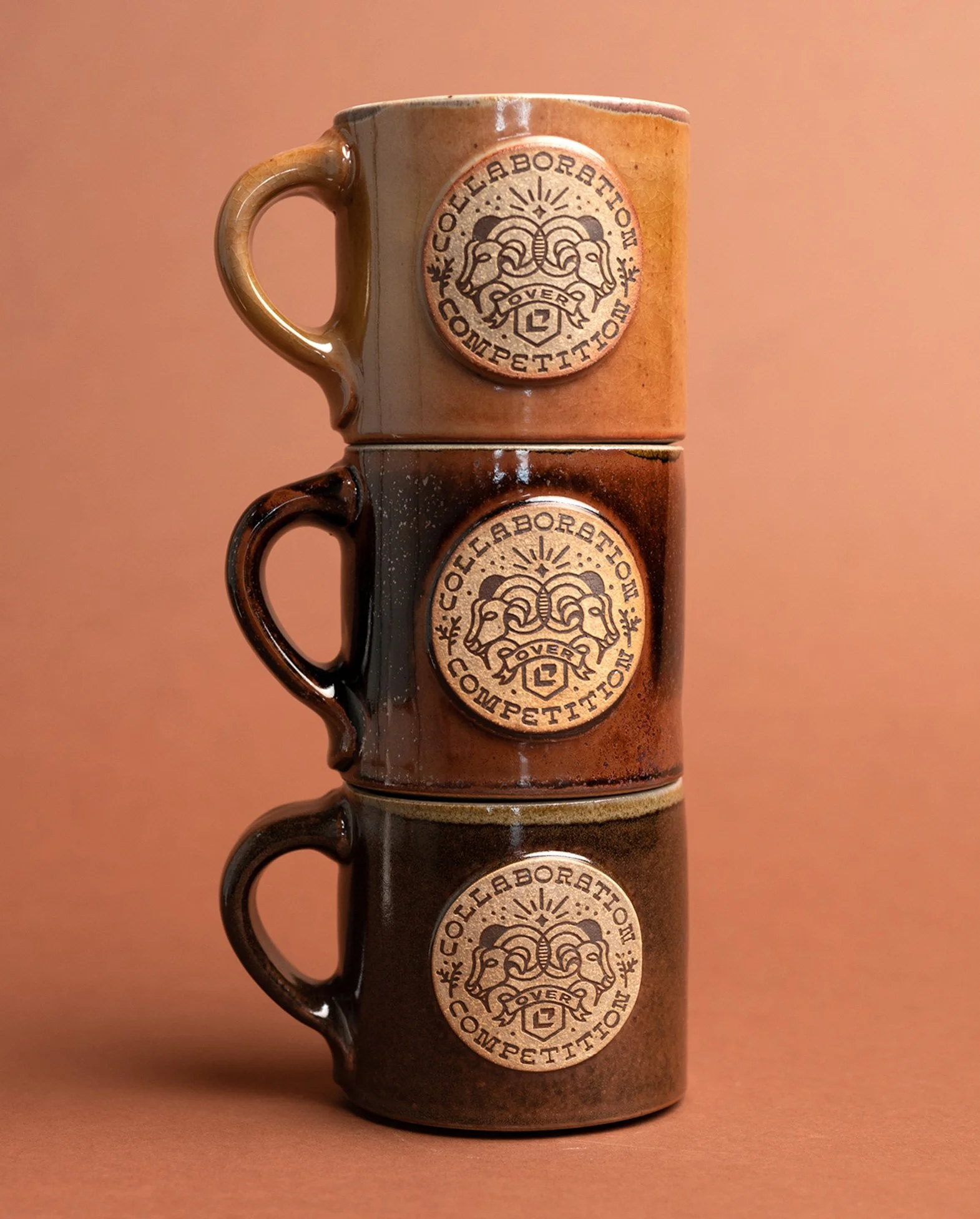 Stack of three ceramic coffee mugs with a logo of two ram's heads and the text 'Collegeborah Over Competition' on each mug, on a brown background.