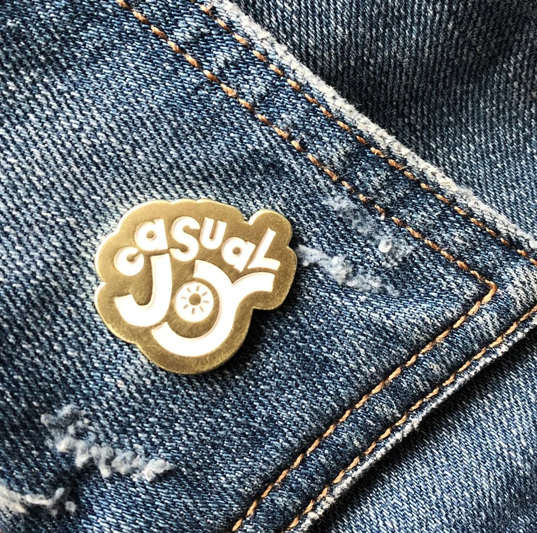 Close-up of a denim fabric with orange stitching and a metallic pin that says 'Joy' with the word upside down, attached to a pocket.