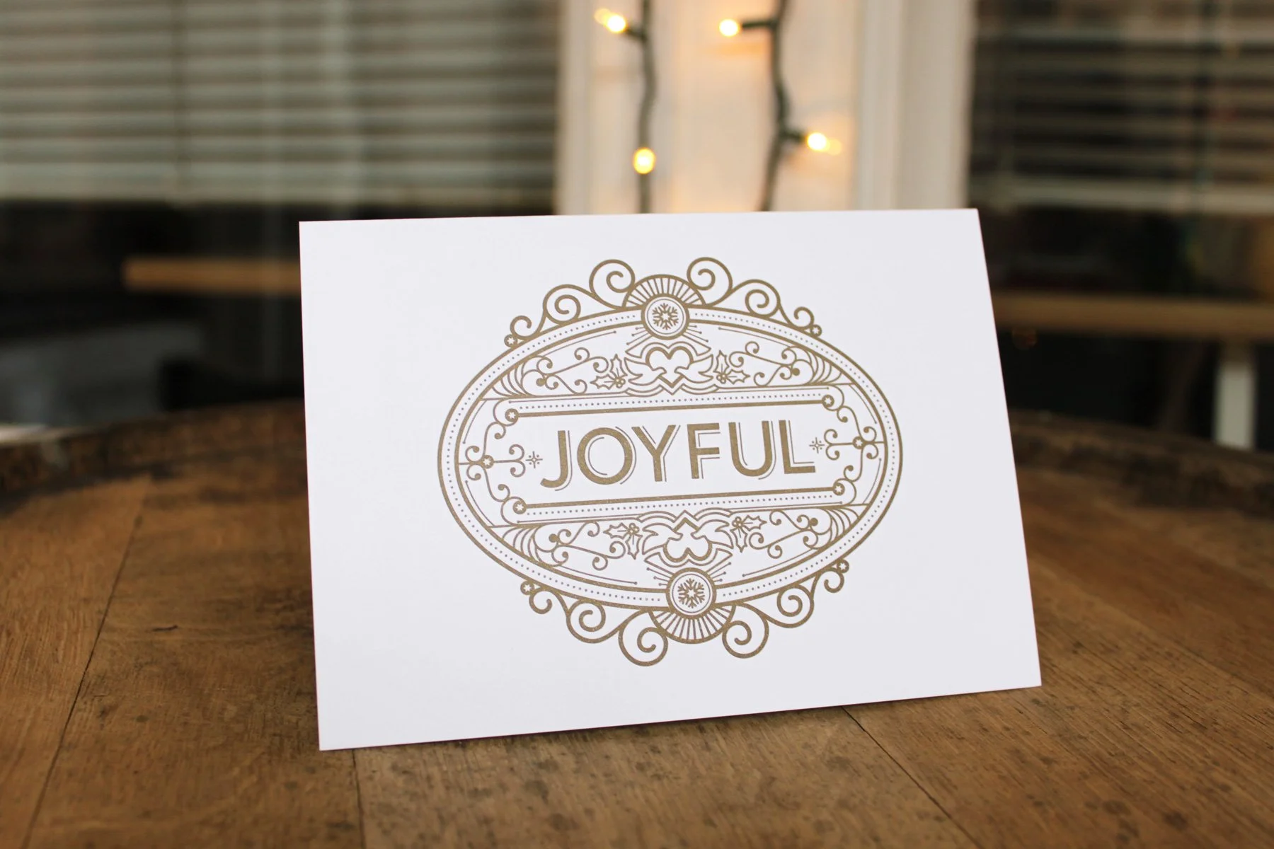 White greeting card with the word "JOYFUL" in decorative gold design, placed on a wooden table with a background of festive lights and window blinds.