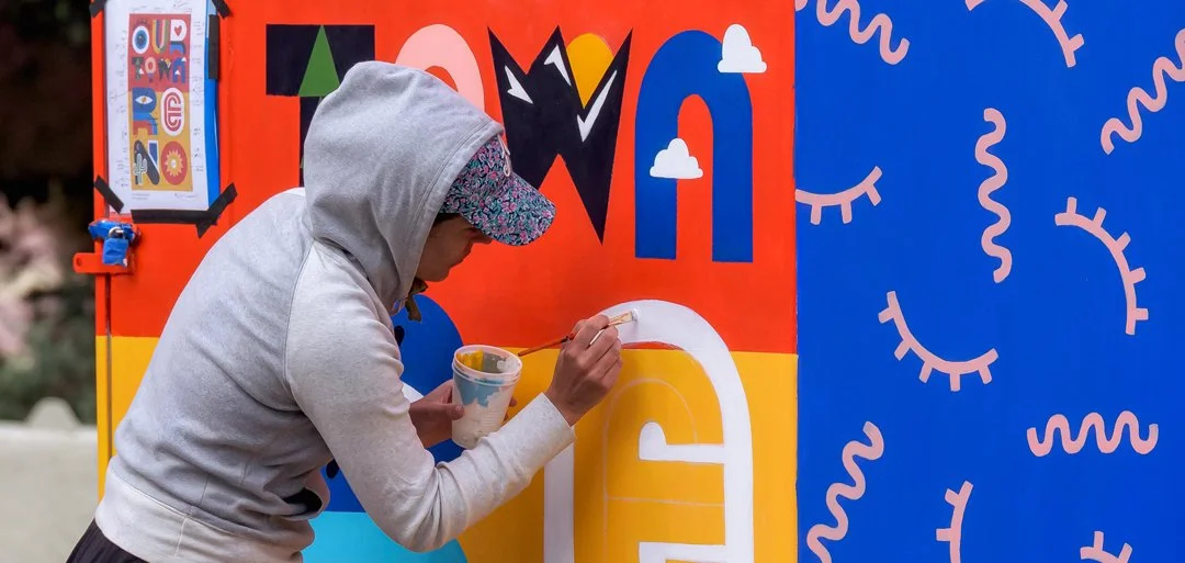 Person painting a colorful mural on a wall, with sections of orange, blue, and yellow, including stylized eyes and clouds in the artwork.