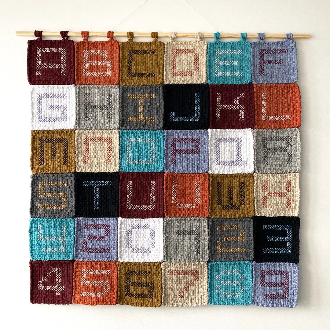 A colorful wall hanging of 30 small quilted squares with numbers and letters stitched in cross-stitch, arranged in a grid on a white wall, each square attached to a wooden dowel.