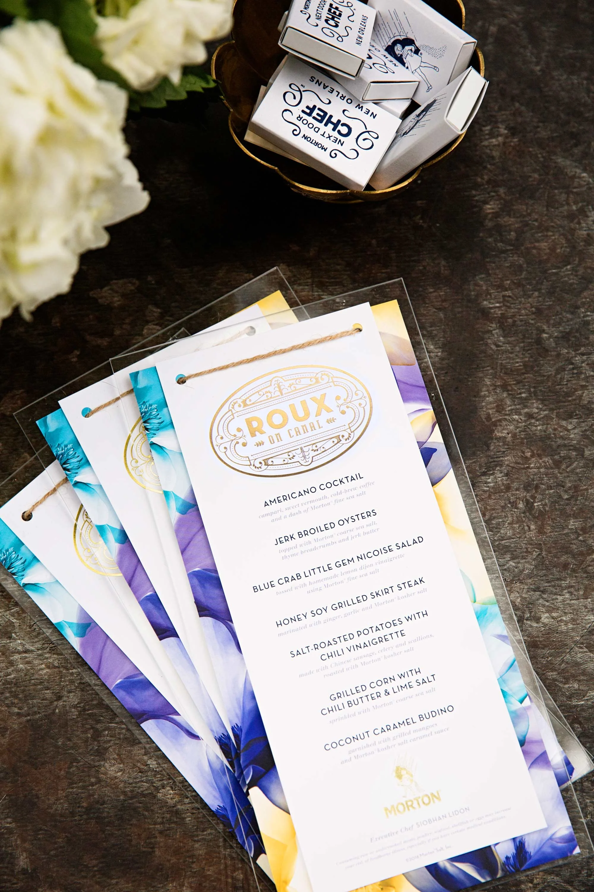 A menu for a restaurant called Roux on Canal, listing dishes such as American Cocktail, Jerk Broiled Oysters, Blue Crab Little Gem Nicoise Salad, Honey Soy Grilled Skirt Steak, Salt-Roasted Potatoes with Chili Vinegarette, Grilled Corn with Chili Butter & Lime Salt, and Coconut Caramel Budino, placed on a dark wooden table. In the background, there are small boxes labeled 'Morton', and some white flowers are partially visible.