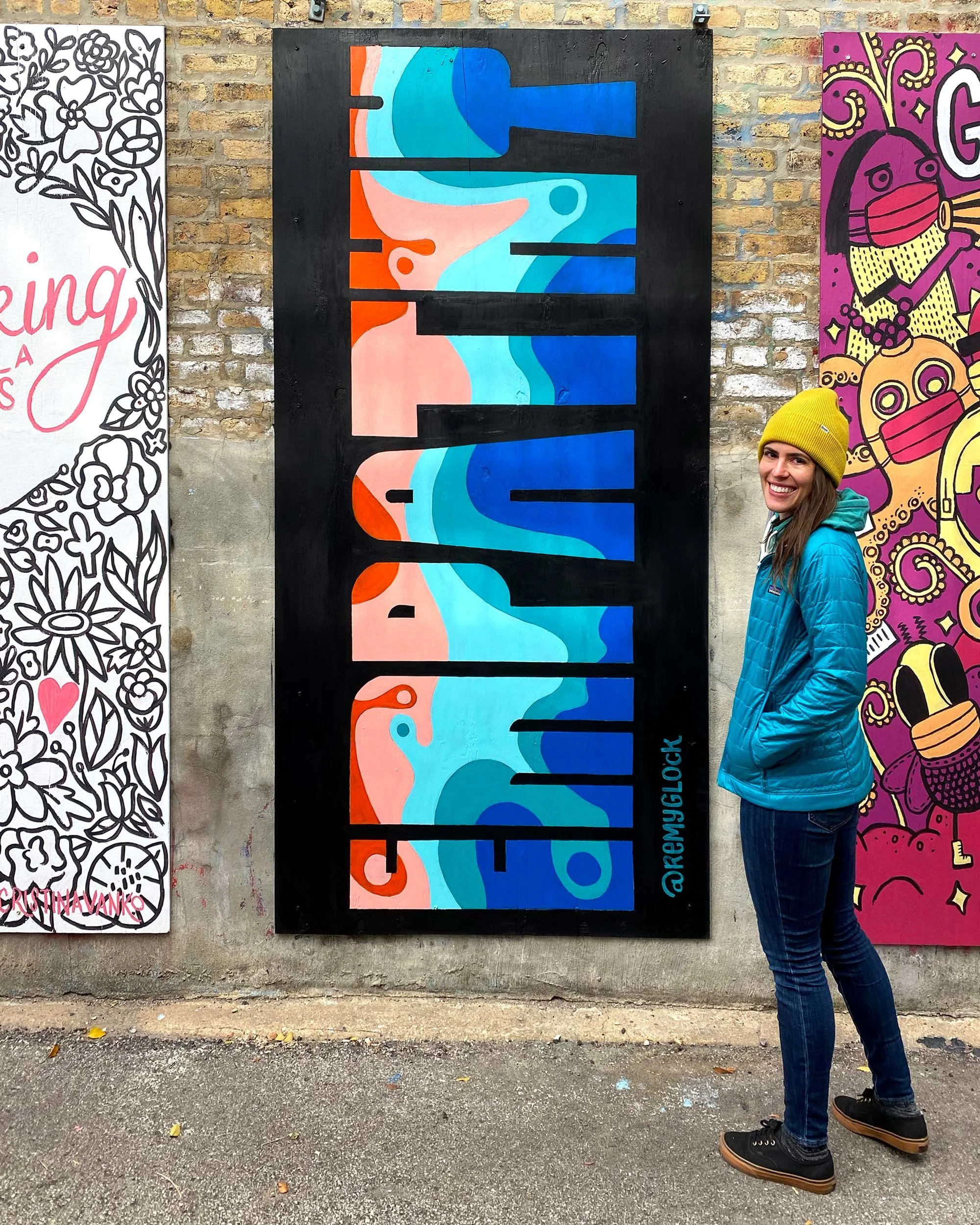 A woman wearing a yellow beanie and blue jacket smiling while standing in front of colorful street art murals on a brick wall. The central mural spells out 'RELAX' with abstract blue and orange shapes.