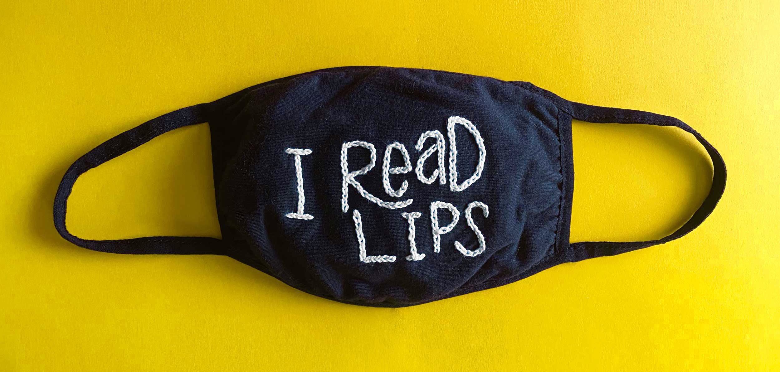 Black face mask with white embroidery that says 'I READ LIPS' on a yellow background