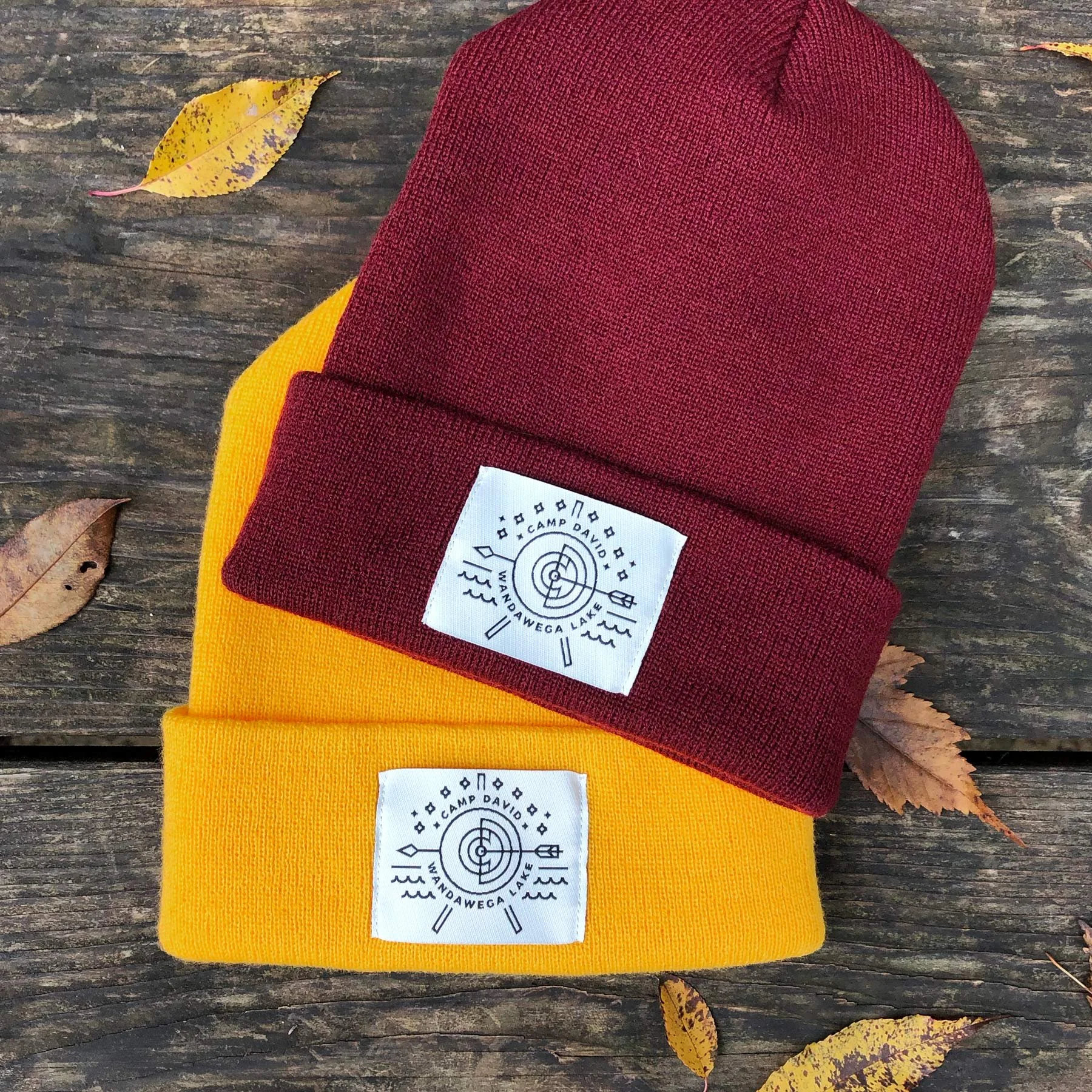 Two knit beanies, one maroon and one yellow, with labels featuring a target, water, and trees, laid on a weathered wooden surface with scattered yellow and brown autumn leaves.
