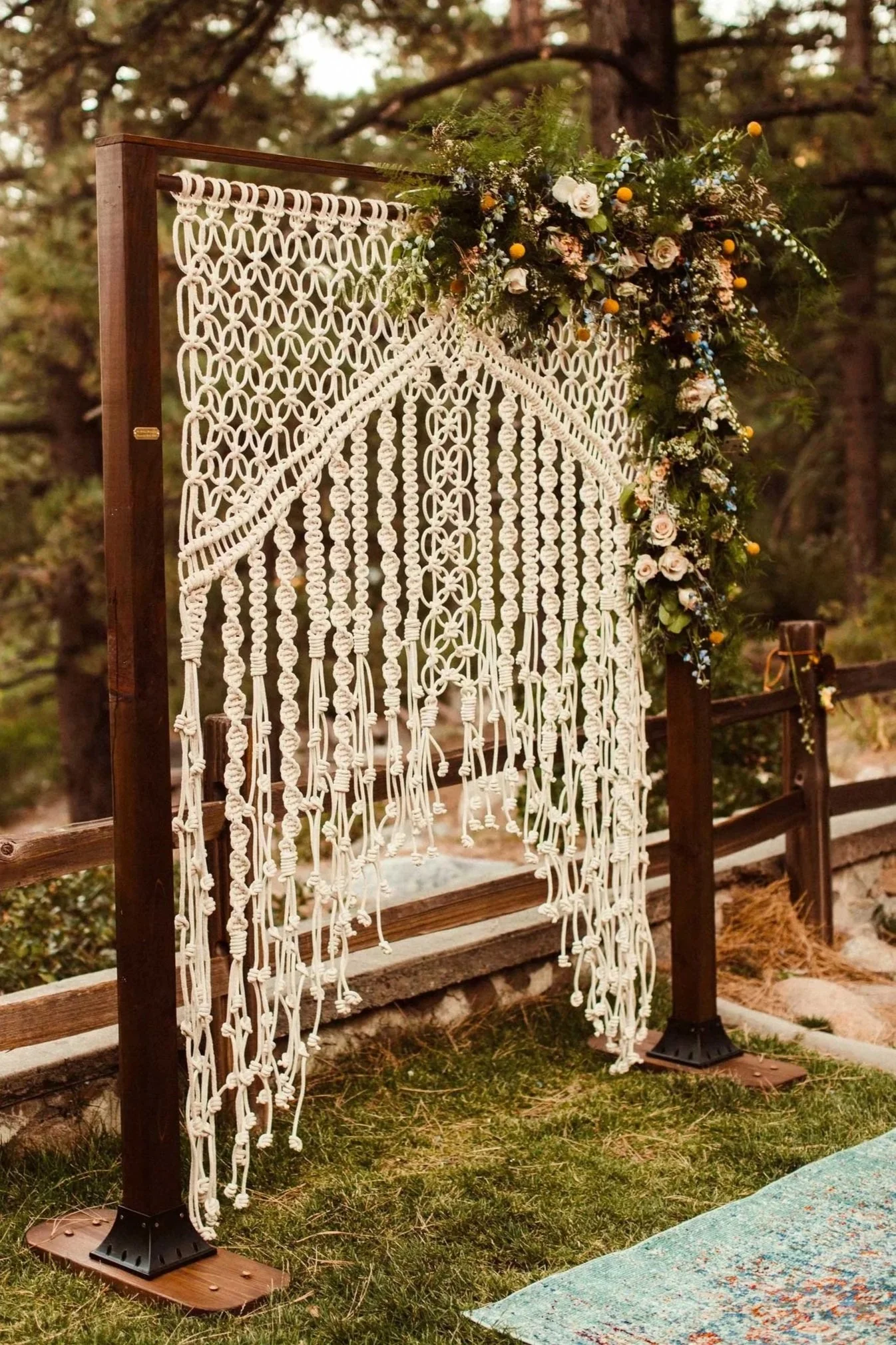 A decorative wedding arch with a macrame backdrop and a floral arrangement of roses, greenery, and small yellow and blue flowers, set outdoors against a forest background.