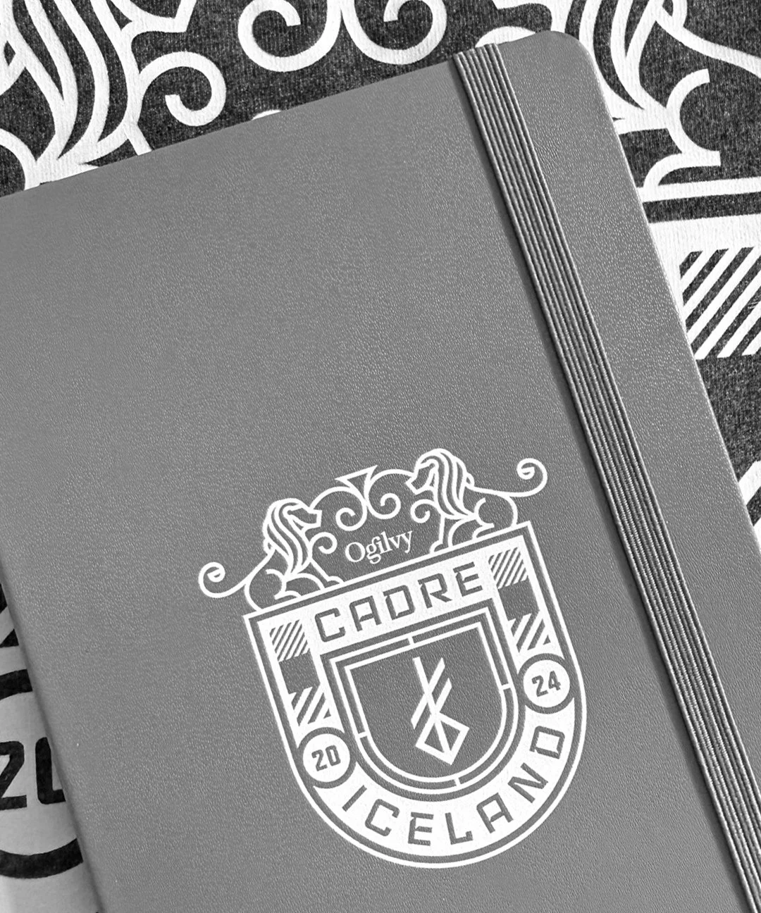 Gray notebook with a logo that reads 'Oglvy, Caure, Iceランド' featuring a shield, a clock, and decorative elements, on patterned background.