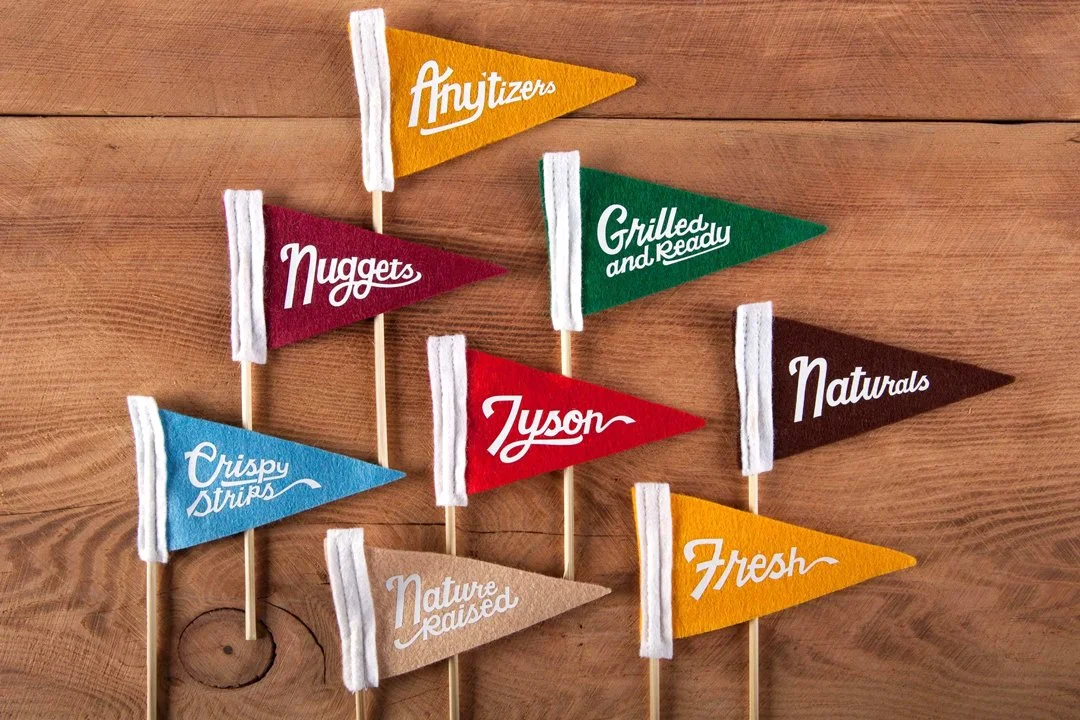 Several small colorful flags with white stripes on sticks placed on a wooden surface. The flags have text, including 'Any Titers,' 'Nuggets,' 'Grilled and Ready,' 'Naturals,' 'Crispy Strips,' 'Tyson,' 'Nature Raised,' and 'Fresh.'