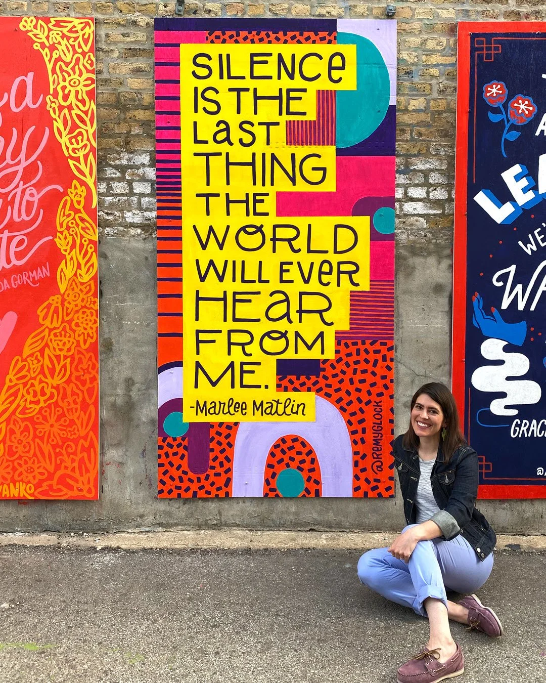 Colorful mural graffiti with a quote by Maudee Matlin that says, 'Silence is the last thing the world will ever hear from me,' located on a brick wall, with a woman kneeling and smiling in front of it.