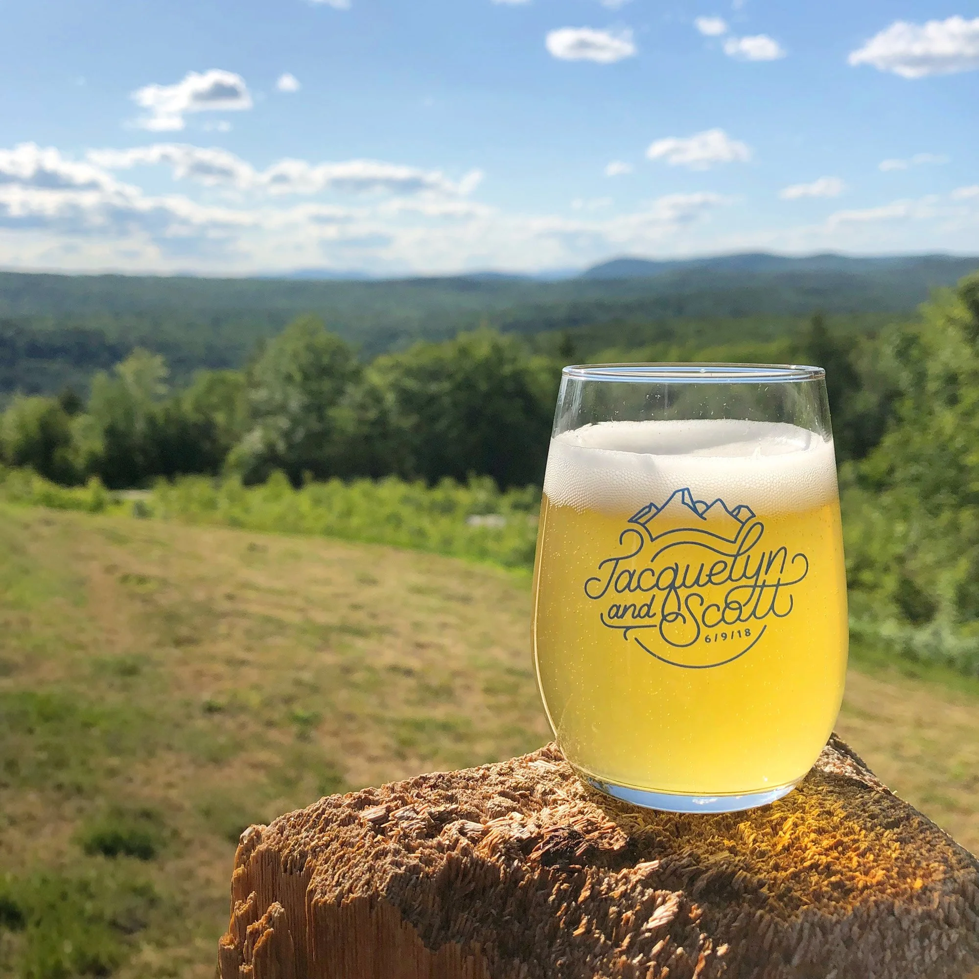 A glass of beer with a personalized wedding or event label reading 'Jacquelyn and Scott 6/9/18' placed on a wooden surface outdoors, with a scenic mountain and forest landscape in the background under a partly cloudy sky.