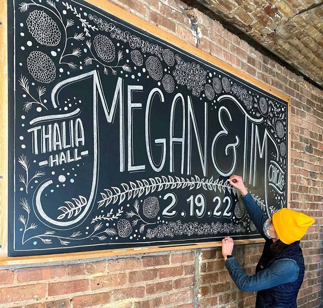 A person wearing a yellow beanie is drawing or writing on a large black chalkboard with white and blue chalk. The chalkboard features decorative floral and geometric designs around the text 'Megan & Jim' and information about an event happening on February 19, 2022, at Thalia Hall in Chicago.