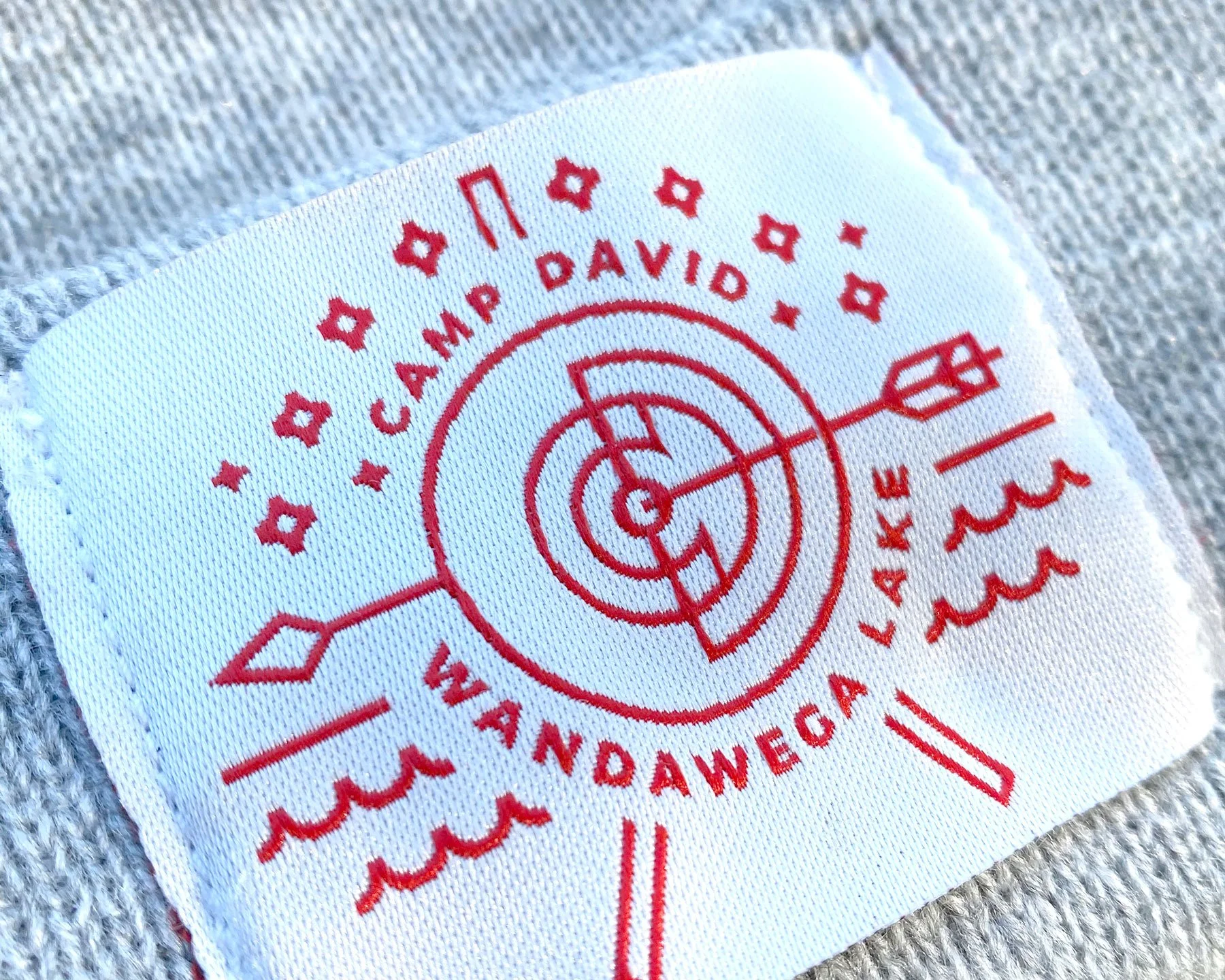 Close-up of a white fabric patch with red printed design featuring the name 'CAMP DAVID' and the word 'LAKE' with wavy lines, directional arrows, a compass rose, and decorative stars surrounding a central circular pattern.