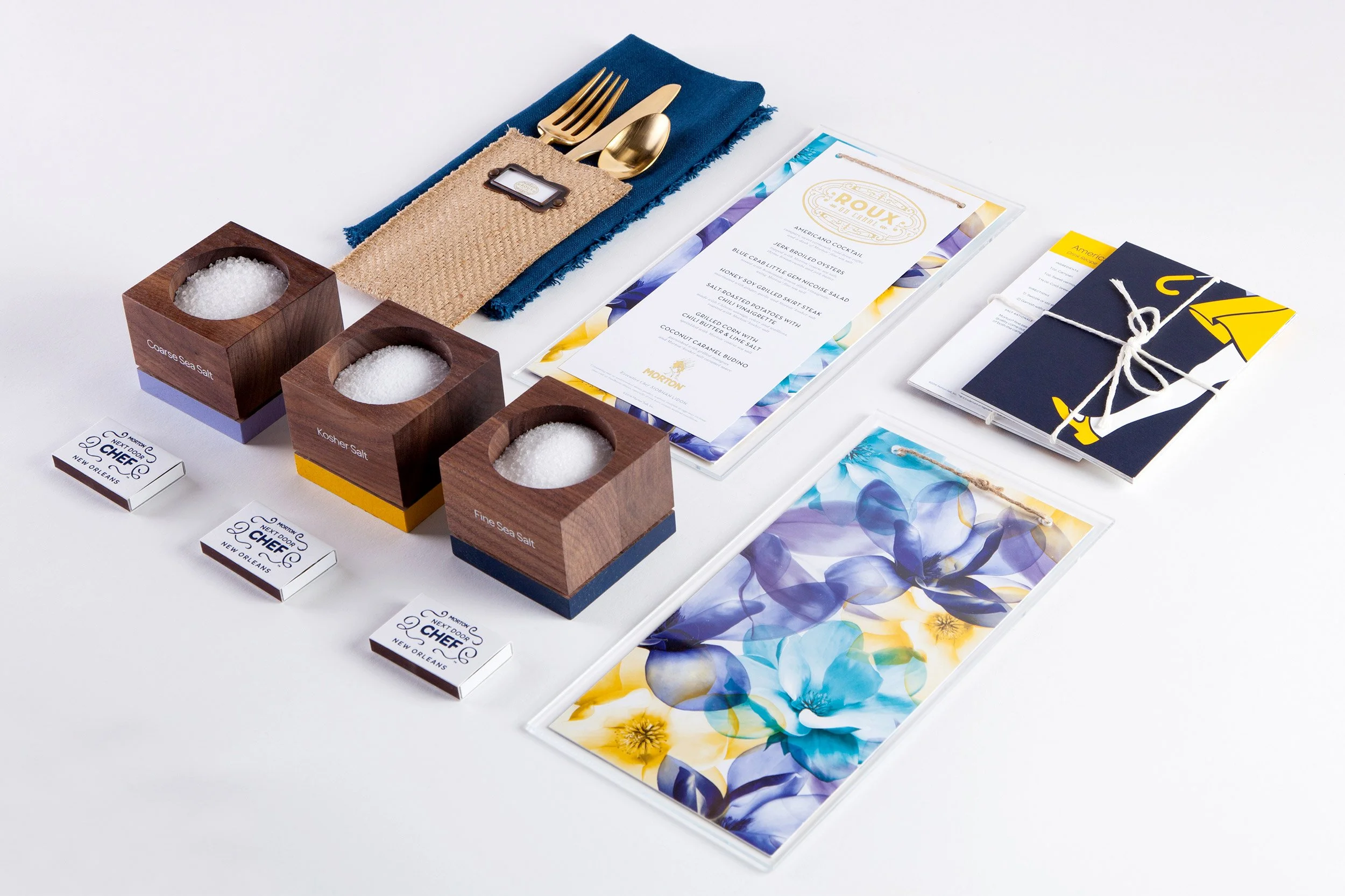 A table setup with three boxes of different types of sea salt, black, kosher, and fine, along with small containers and a menu, a set of utensils wrapped in fabric, and printed materials tied and laid out on a white surface.