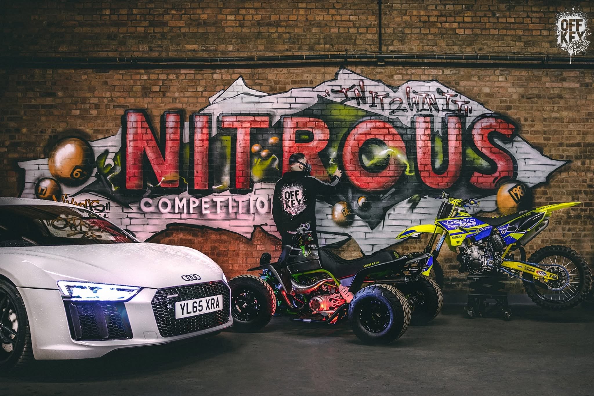 A graffiti mural on a brick wall featuring the words 'Nitrous Competition' in large, red letters with a mountain illustration in the background. In front, there is a white Audi car, a person wearing a black 'OFF KEY' jacket using a spray can to paint, and two dirt bikes, one yellow and blue and the other black and red, with the bikes and person positioned in front of the mural.