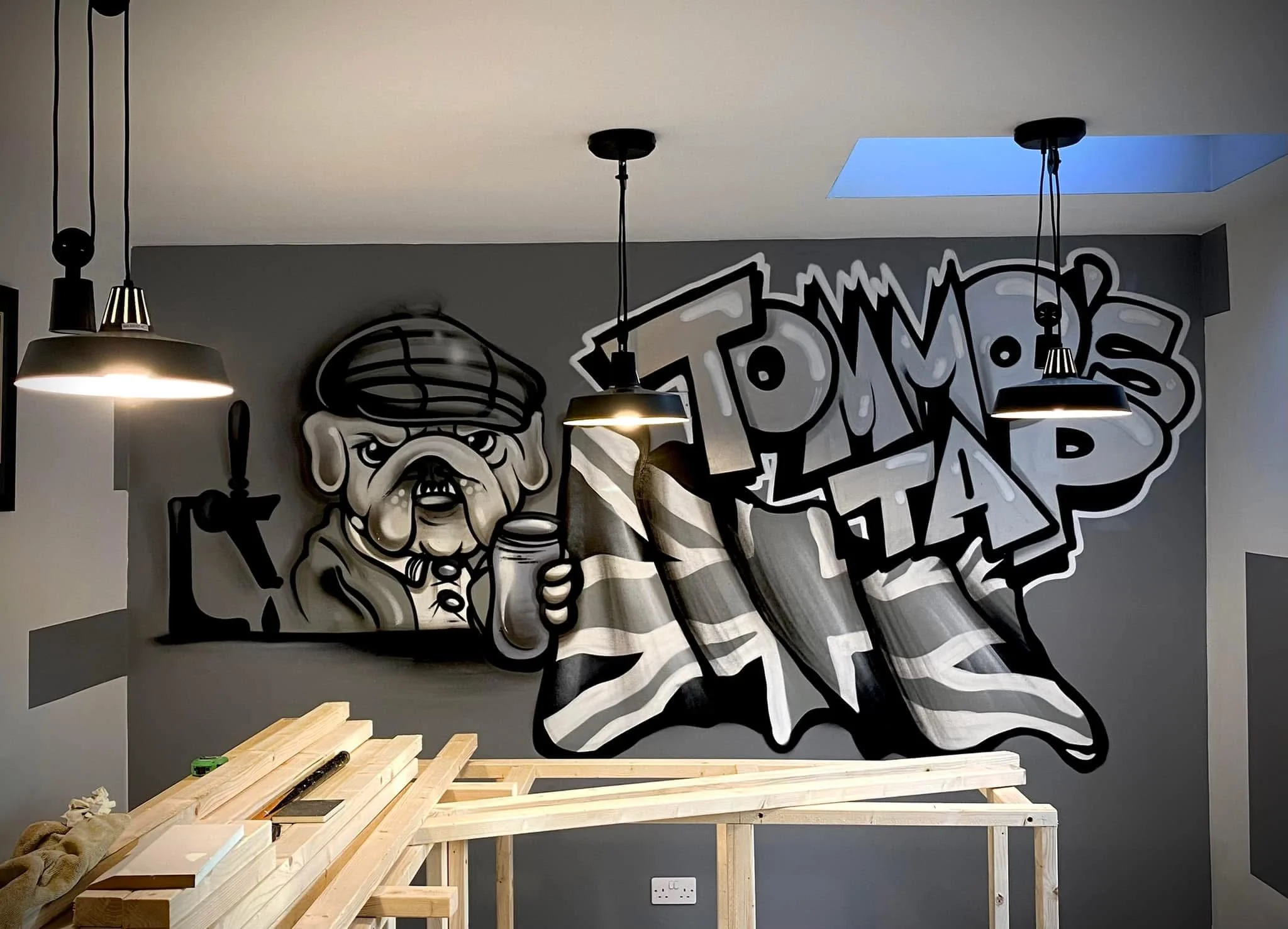 Graffiti wall art of an elderly man with a beard and hat, holding a jar, next to stylized graffiti text reading "TOMMY'S TAP" in black, white, and gray tones.
