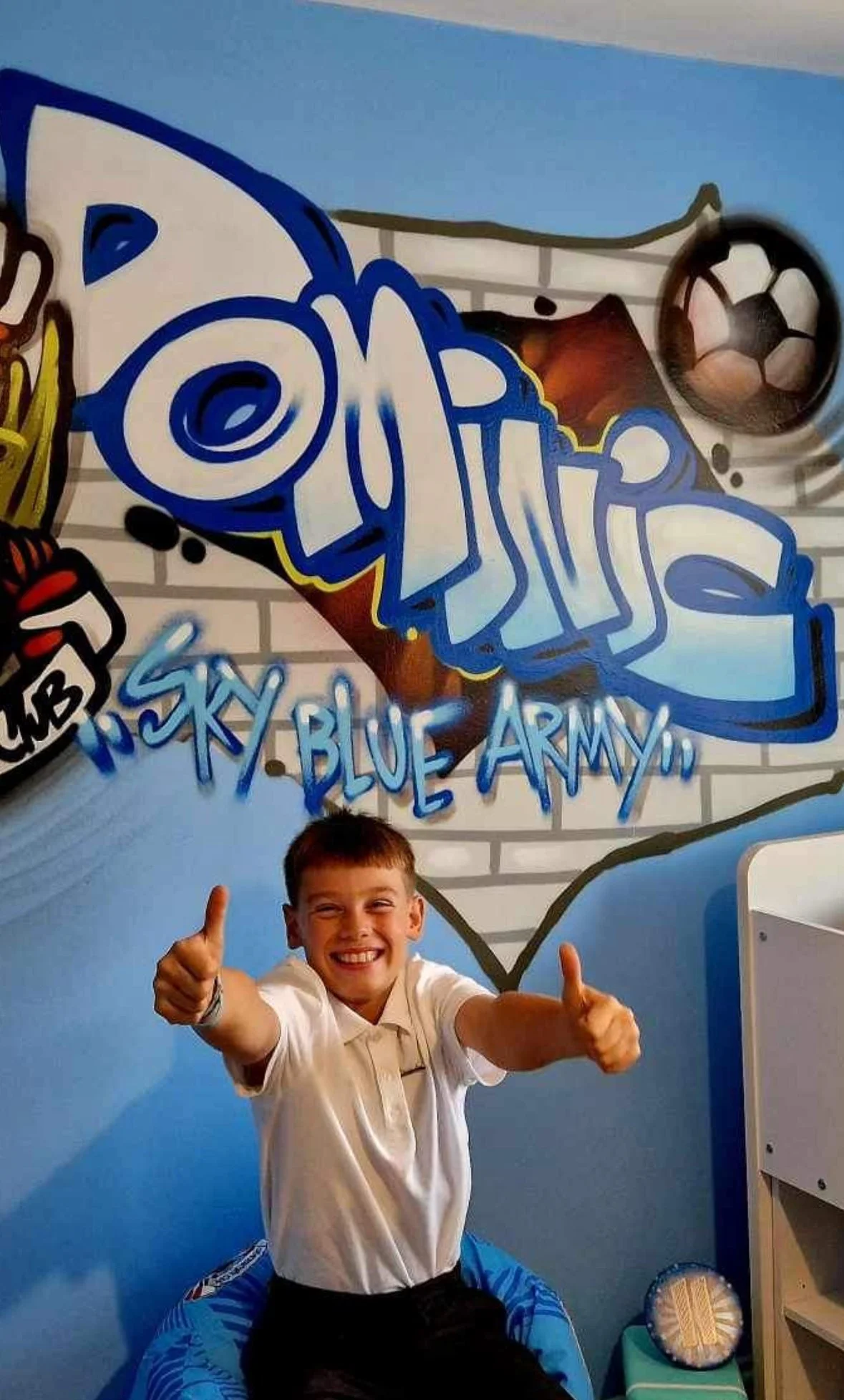 A boy sitting on a blue chair, smiling and giving two thumbs up in front of a graffiti mural with the words Sky Blue Army, wearing a white polo shirt in a room decorated with sports-themed graffiti and a soccer ball image.