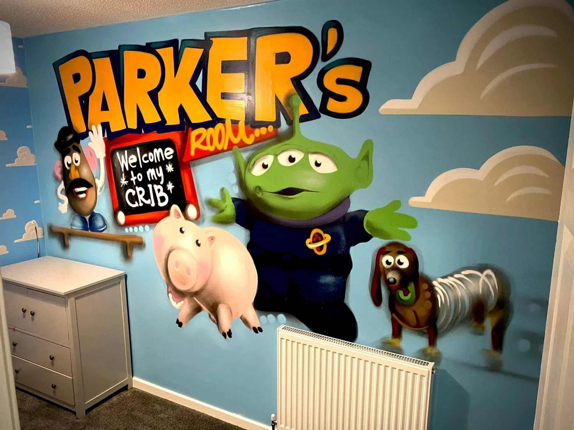 Colorful mural of characters from Toy Story, including Buzz Lightyear dressed as an alien and a pig, on a blue wall with clouds, with a welcome sign and the words 'Parker's Rool...'