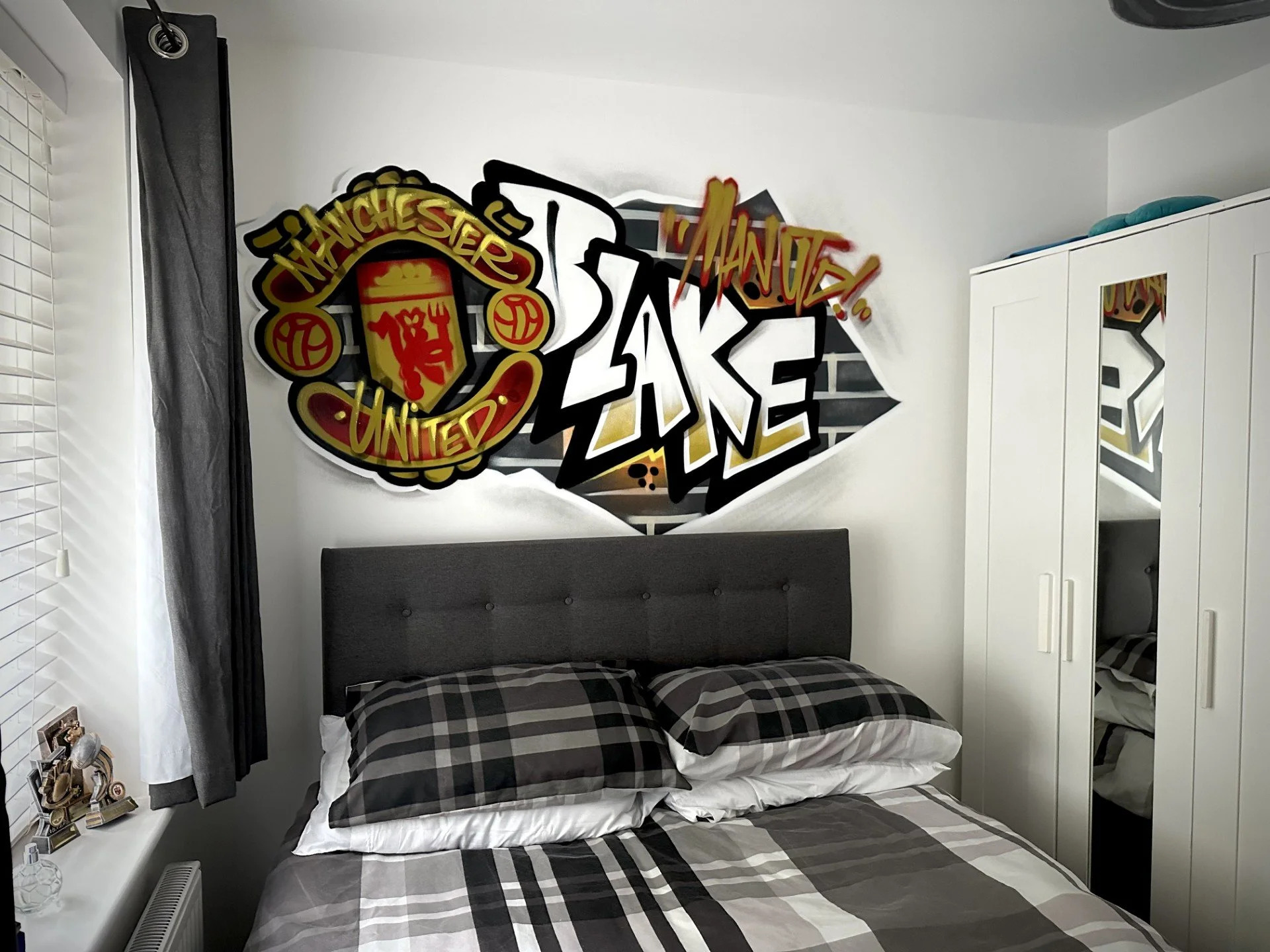 A bedroom with a large graffiti-style Manchester United sign on the wall above the bed, which has a gray headboard and black, white, and gray plaid bedding. There is a white wardrobe with mirrored doors on the right and a window with white blinds and gray curtains on the left.