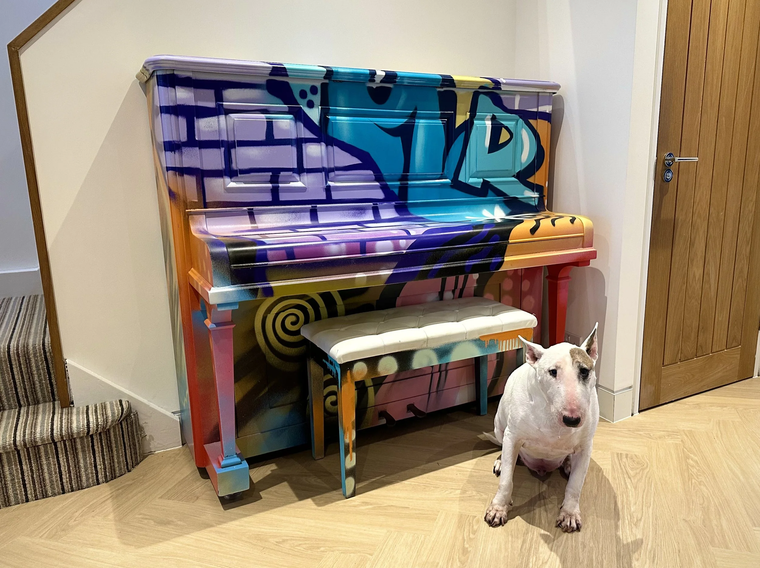 A colorful graffiti-style painted upright piano with a matching bench, positioned in a room with wooden flooring and a dog sitting nearby.