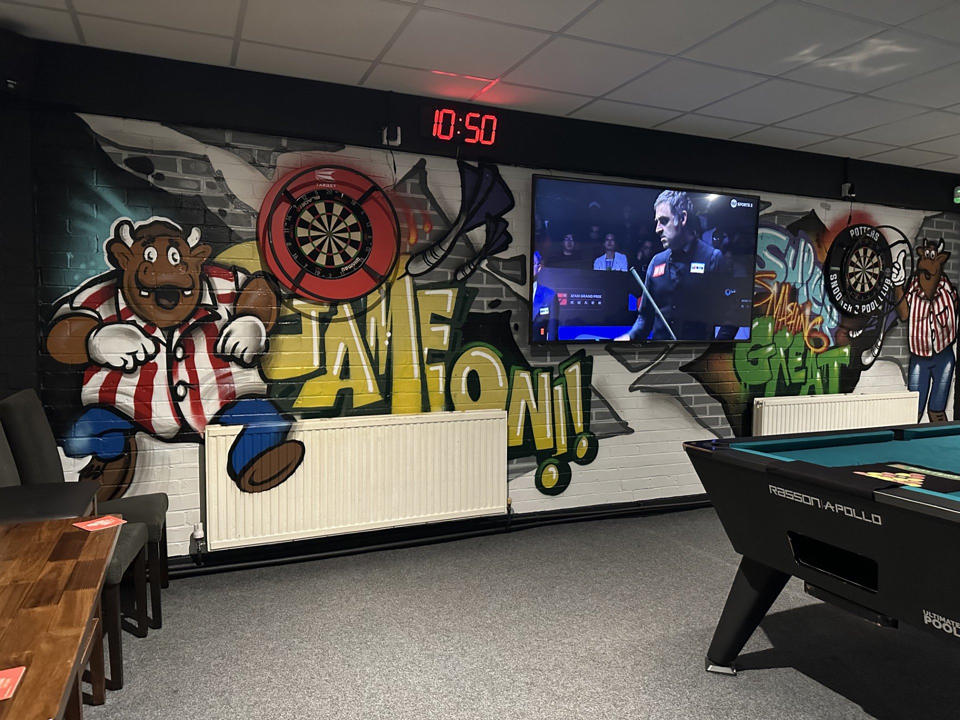 Decorative wall with cartoon bull character in a striped jersey and blue pants, a dartboard, graffiti art, and a flat-screen TV showing a pool tournament, in a game room with a pool table and chairs.