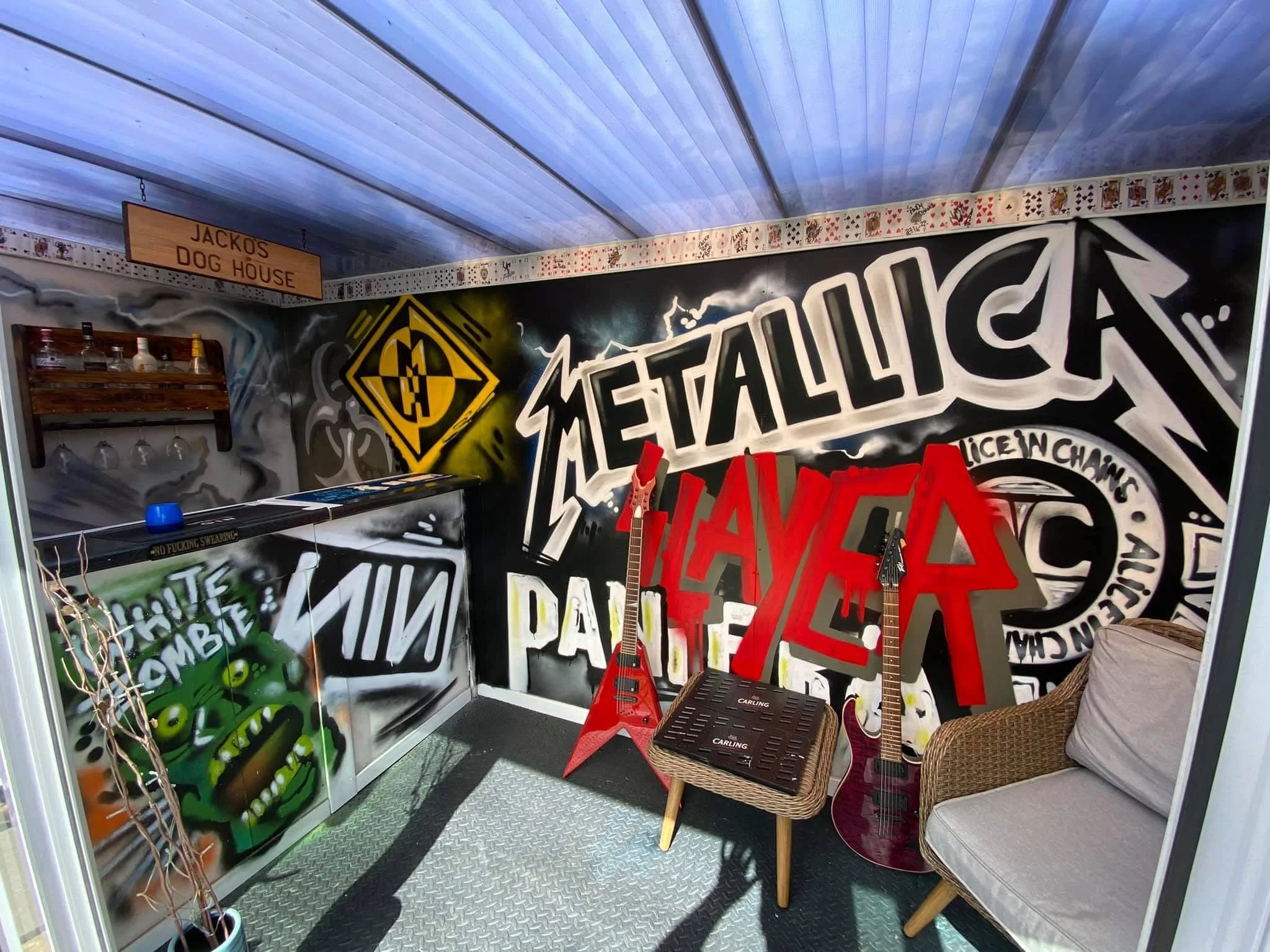 A colorful graffiti-style room with a large wall mural featuring the band Metallica and various artwork. There are two red guitars, a small wicker table, and a cushioned chair. A wooden sign reads 'JACKO'S DOG HOUSE' at the top, and there are some bottles on a shelf on the left side.