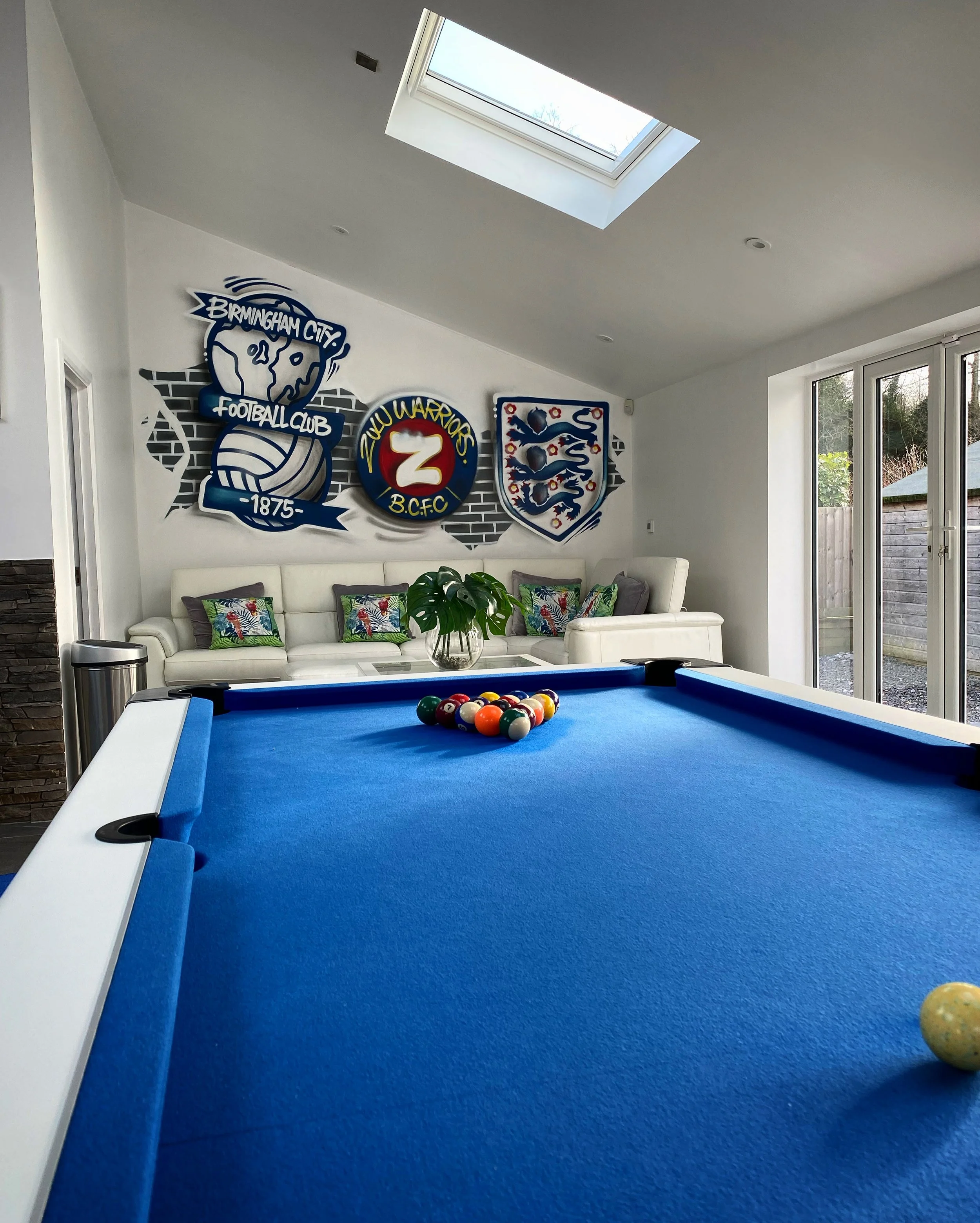 A blue pool table with balls racked in the center, a white sofa with colorful cushions, a potted plant on a white coffee table, and sports team banners on the wall visible in a bright room with skylight and sliding glass doors.