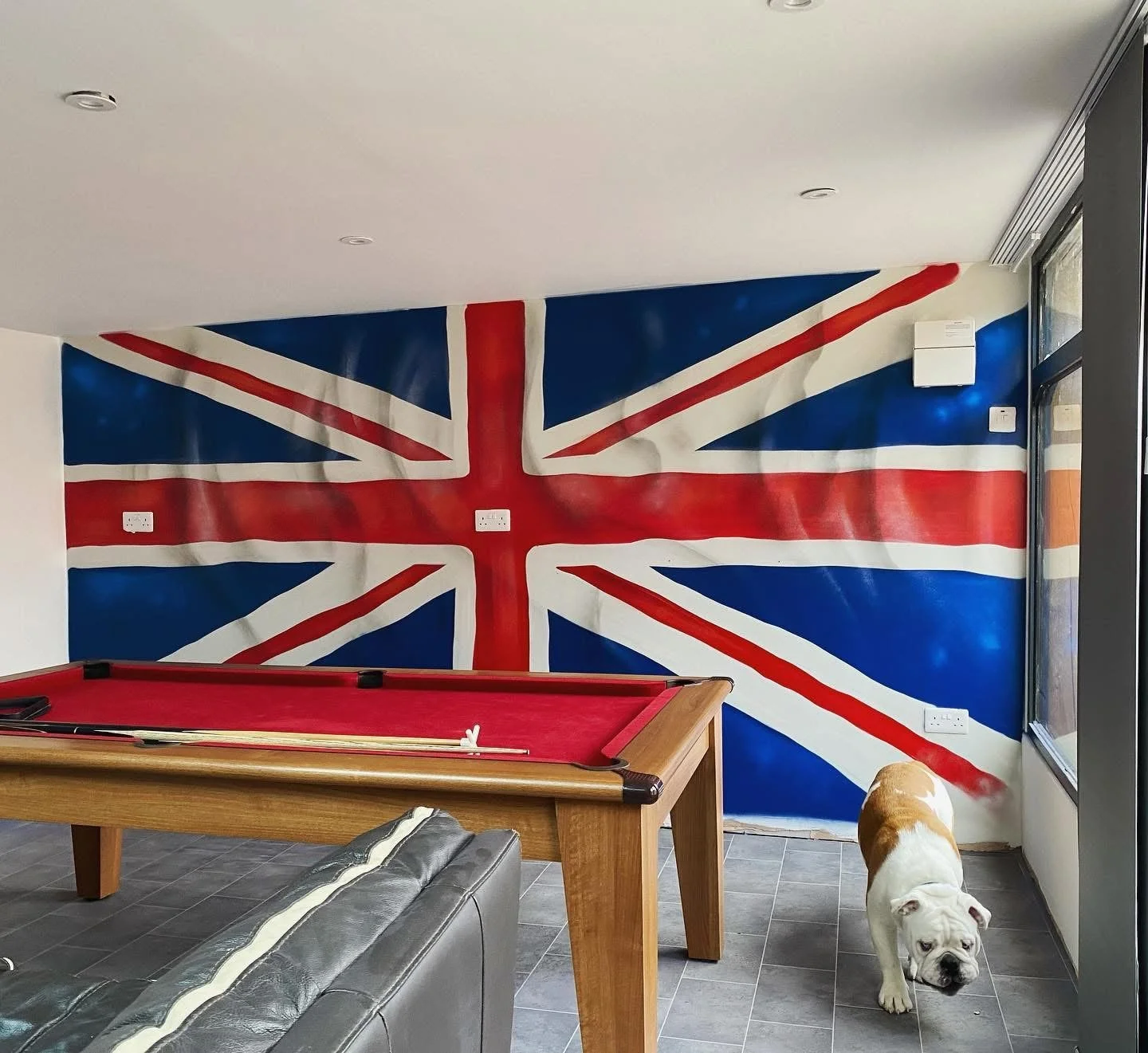 Room with a large Union Jack flag painted on the wall, pool table with red felt, gray leather sofa, and a bulldog walking on tiled floor near a window.