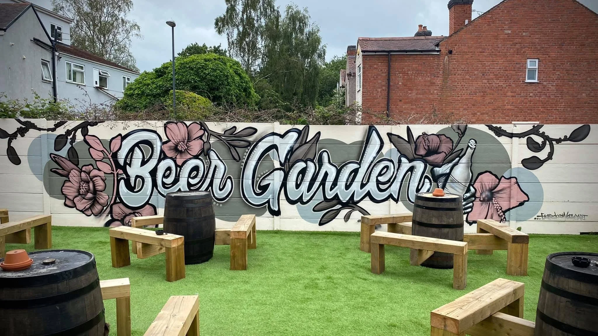 A mural with the words 'Beer Garden' on a white fence, decorated with pink flowers and dark outlines. There are wooden benches and barrels around a grassy area in front of the mural.