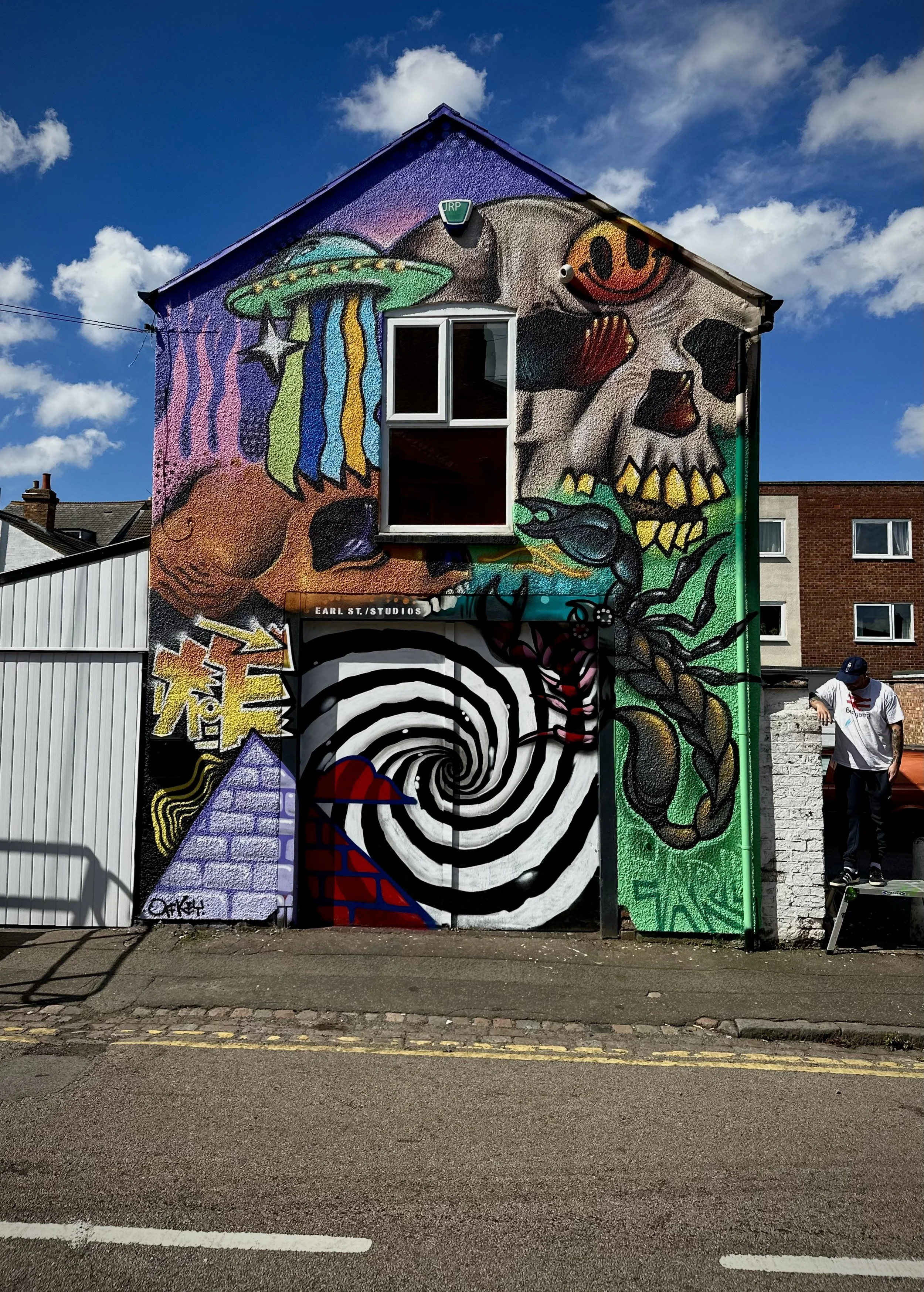 Street art mural on a two-story building featuring a astronaut helmet, a skull with a smiley face, a lobster, and a black and white spiral tunnel with a red and blue pyramid."