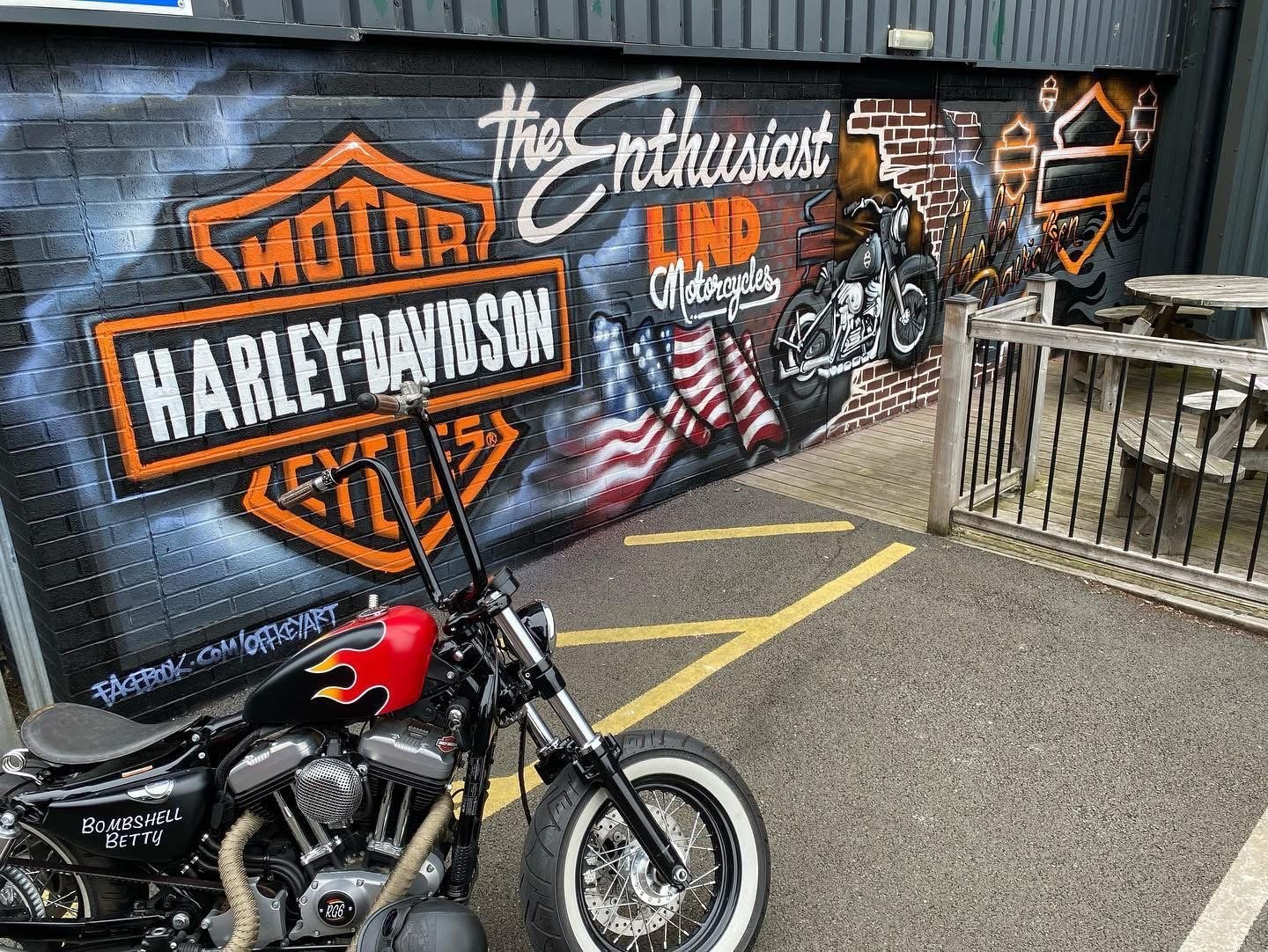A black Harley-Davidson motorcycle with a flame design on the fuel tank and custom exhaust pipes parked in front of a colorful mural. The mural features the Harley-Davidson logo, a motorcycle illustration, an American flag, and various text including "the Enthusiast LAND" and "Harley-Davidson." There is outdoor seating with wooden tables and a fence on the right side.