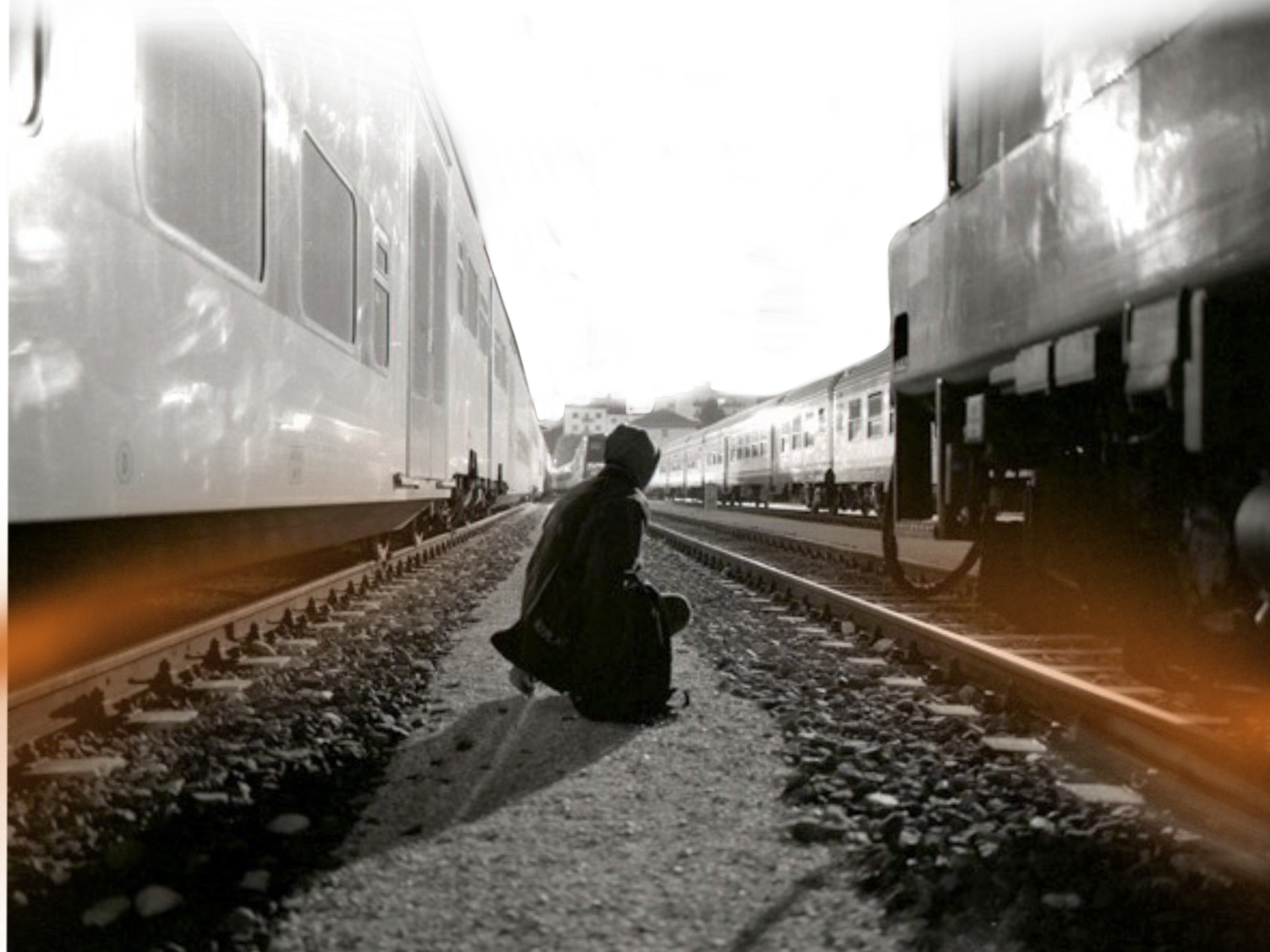 A person squatting on train tracks between two trains at night, with a dark sky and some buildings in the background.