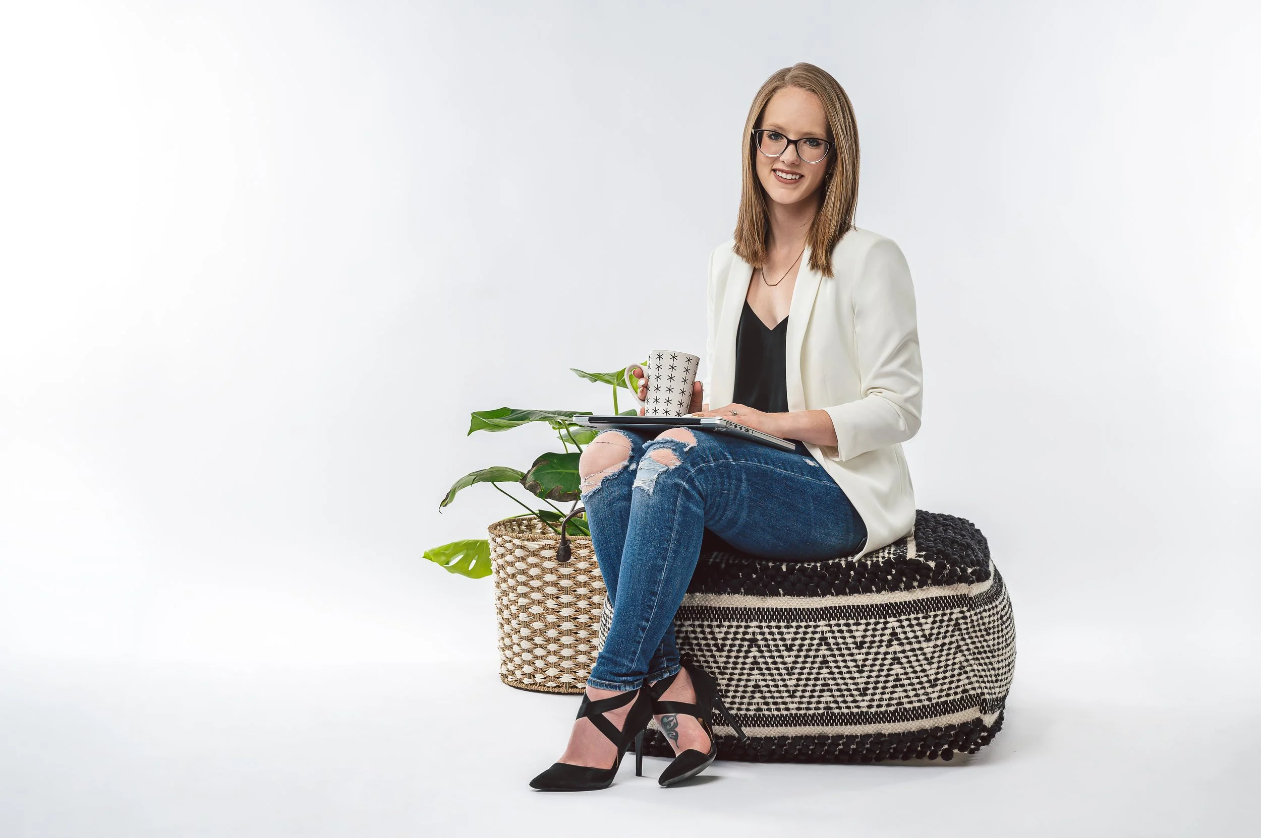 A woman with glasses, wearing a white blazer, black top, ripped jeans, and black high heels, sitting on a textured round cushion with a small plant beside her. She is holding a mug and a laptop, smiling at the camera against a plain white background.