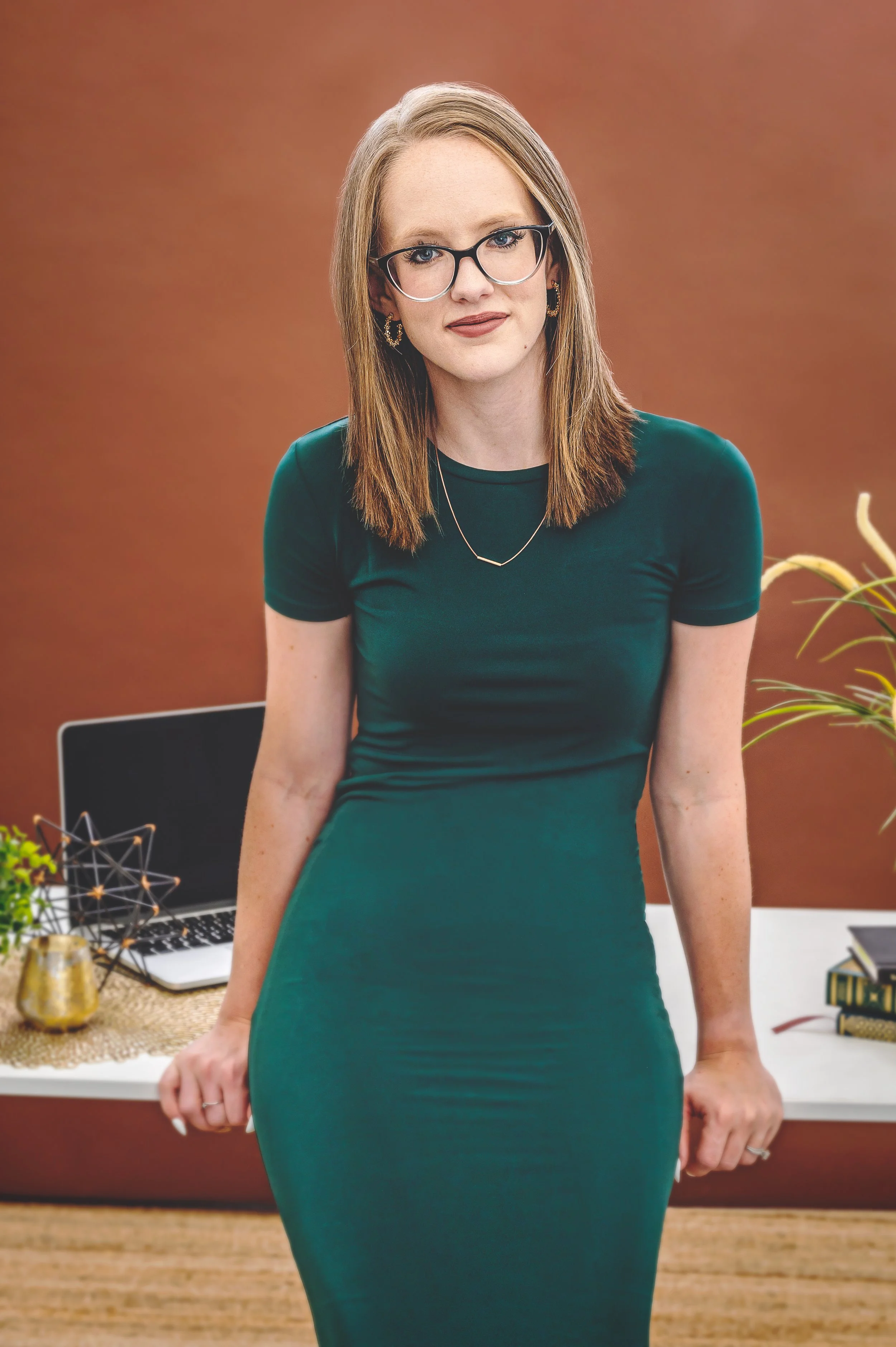 A woman with shoulder-length brown hair, glasses, wearing a emerald green dress, standing indoors with a desk, laptop, and decorative items in the background.