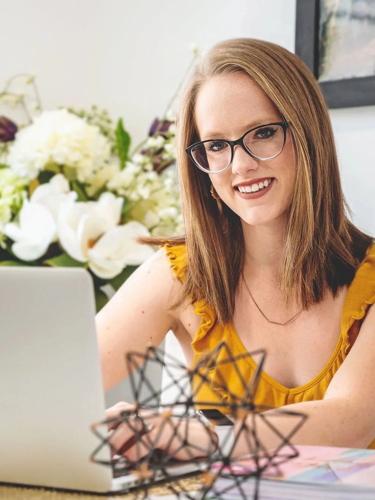 A woman with glasses and straight shoulder-length brown hair smiling at her laptop, with a floral arrangement in the background.