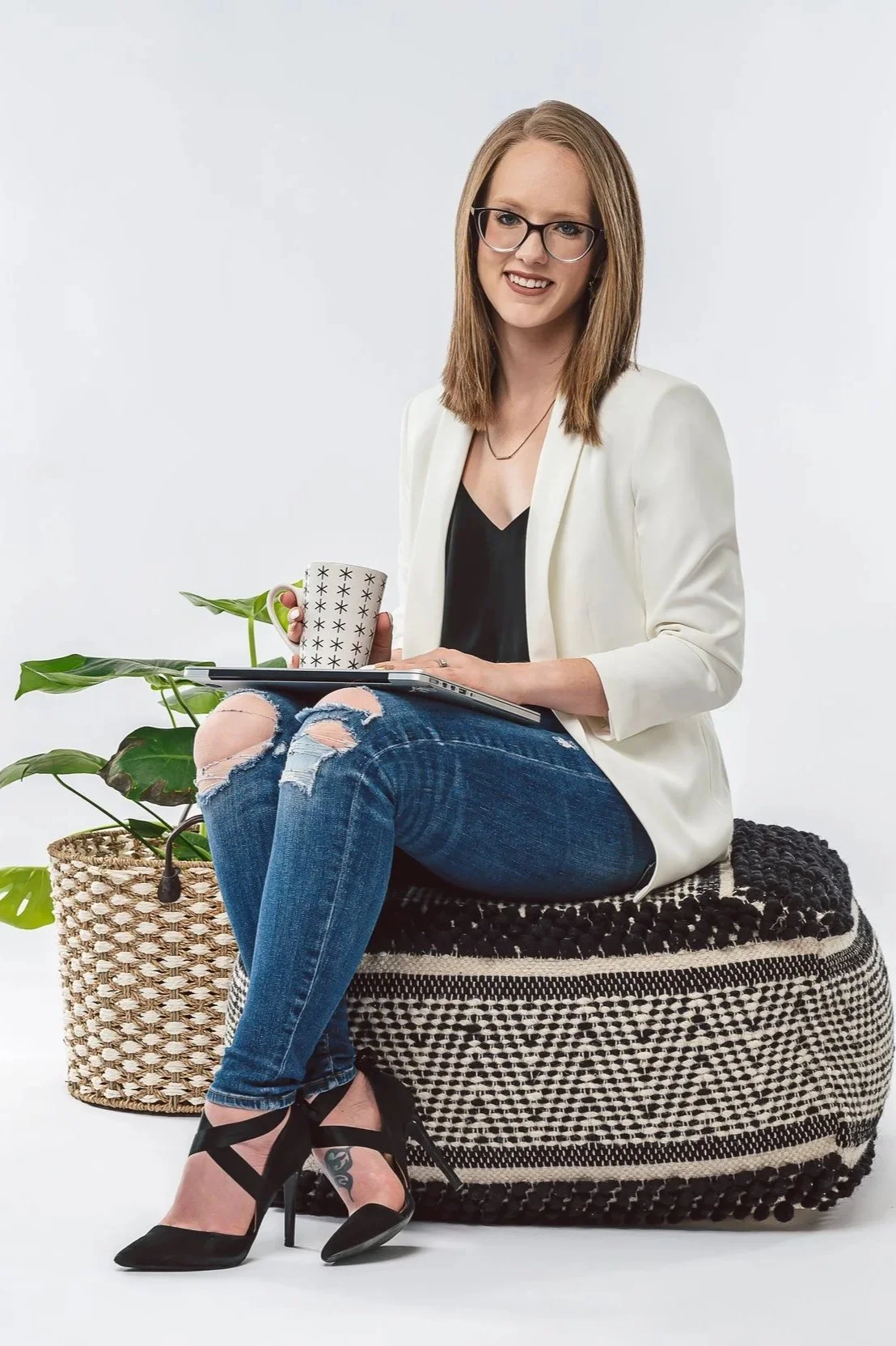 A woman with glasses, wearing a white blazer, black top, and ripped jeans, sitting on a basket with a patterned pouf, holding a coffee mug and a laptop, with a potted plant nearby.