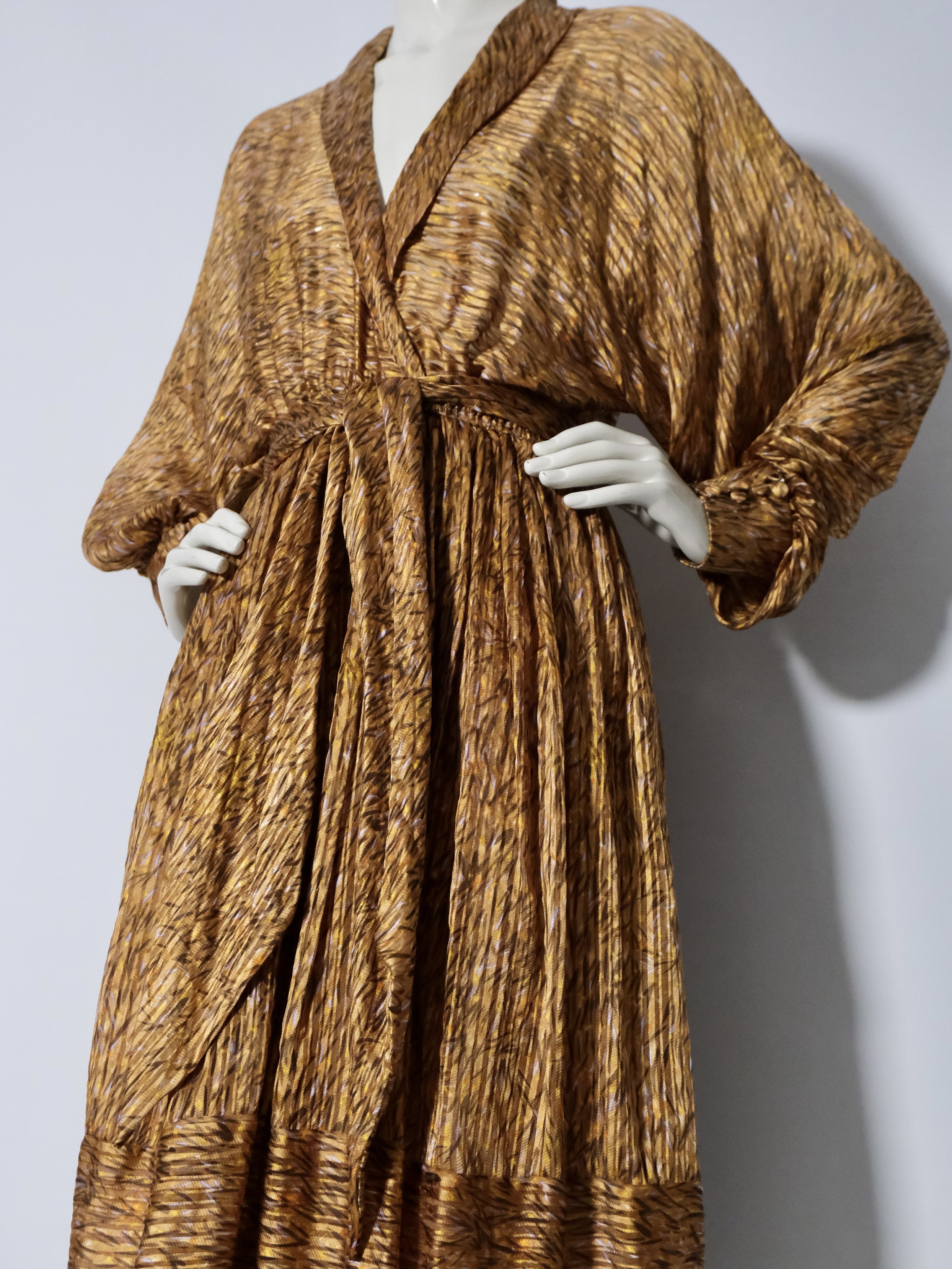A mannequin dressed in a long, gold-colored textured dress with puffed long sleeves, standing against a gray wall.