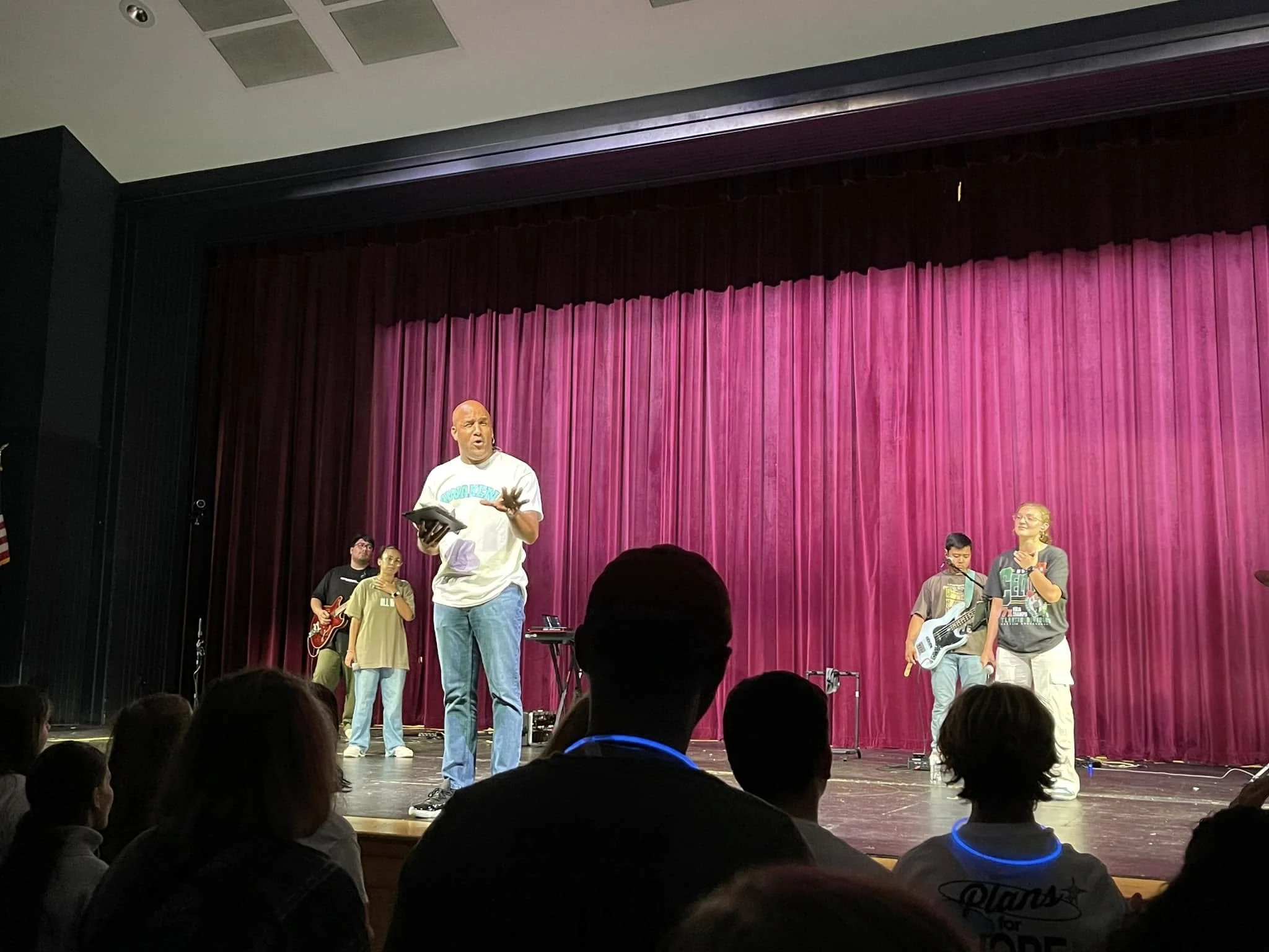 A group of children and a man on stage with a purple curtain, one child is playing guitar, and the man appears to be speaking to the audience.