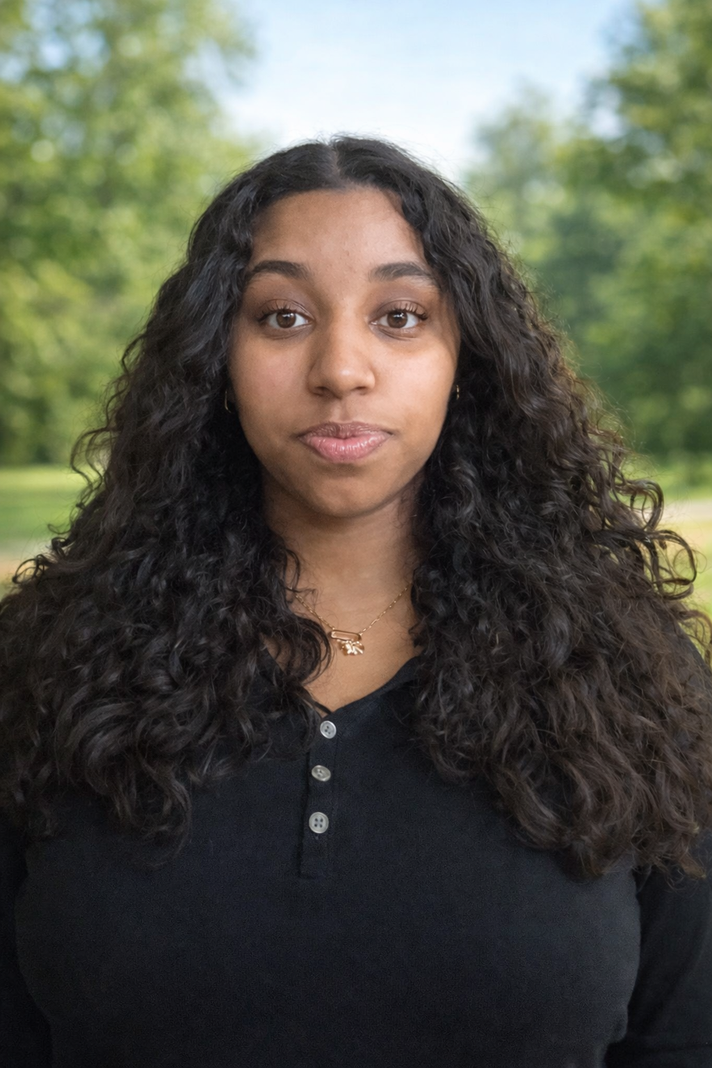Portrait of a young woman with long curly hair, wearing a black top and a gold necklace, standing outdoors with a blurred green park background.
