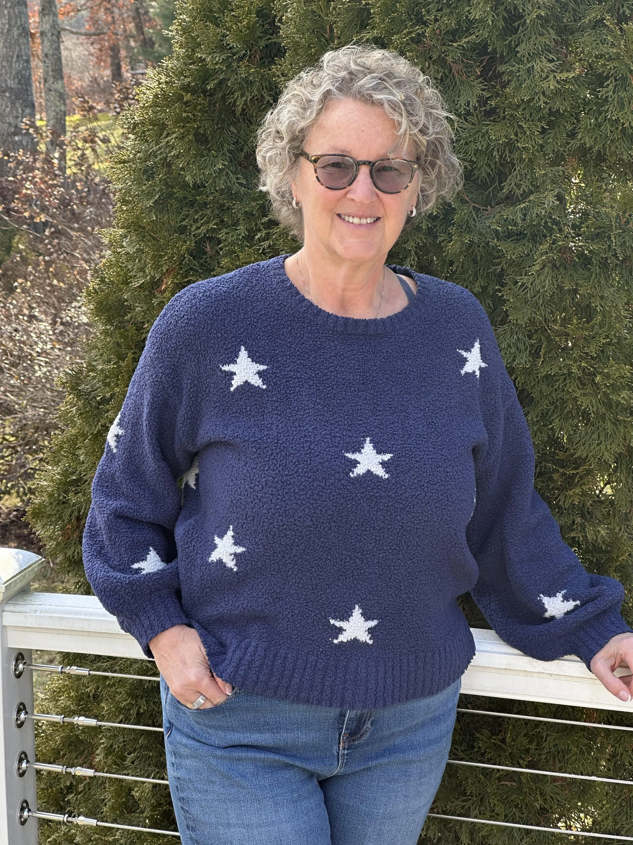 A woman with short curly gray hair and glasses smiling outdoors in front of a large bush, wearing a navy blue sweater with white star patterns and blue jeans.