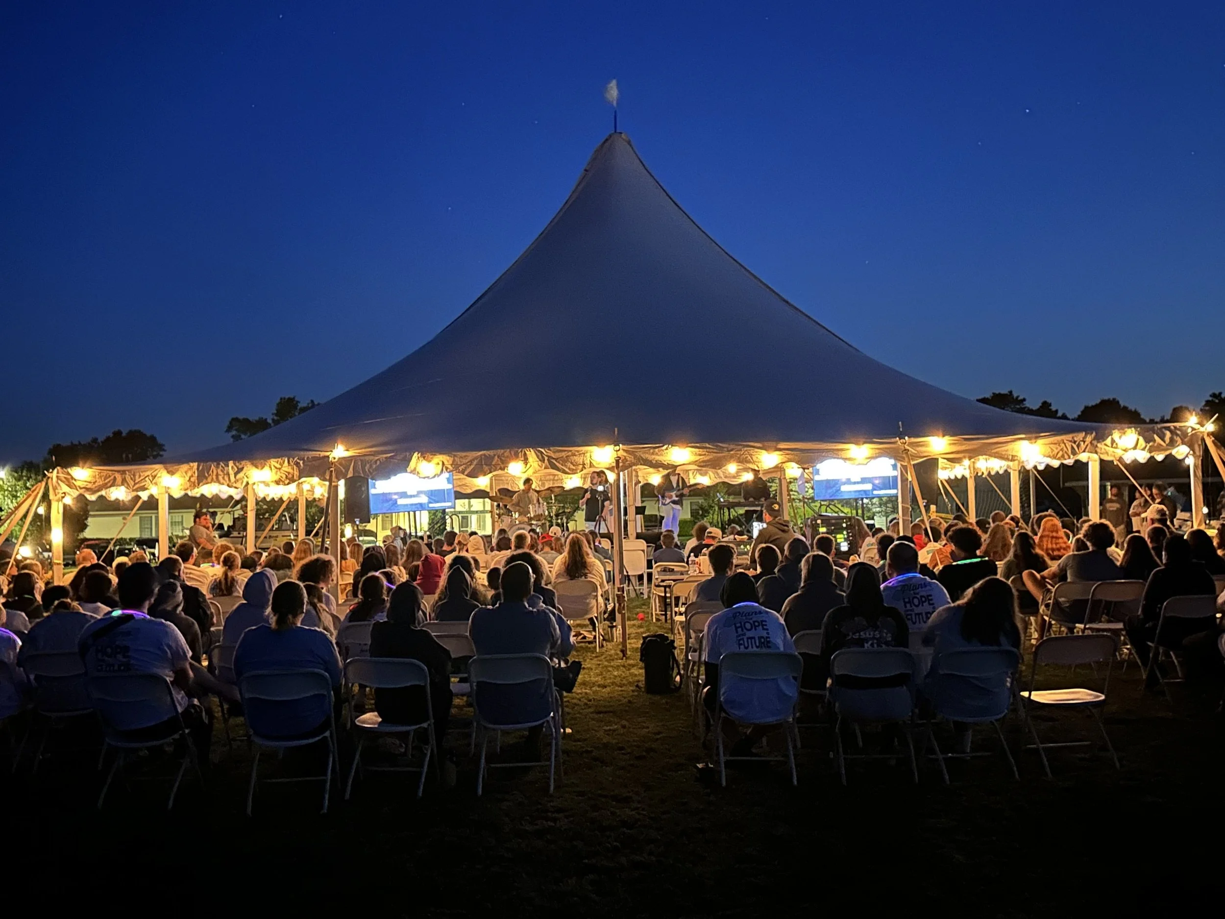 Outdoor evening concert under a large tent with an audience sitting on chairs, a band performing on stage with musical instruments, and string lights hanging from the tent, against a night sky.