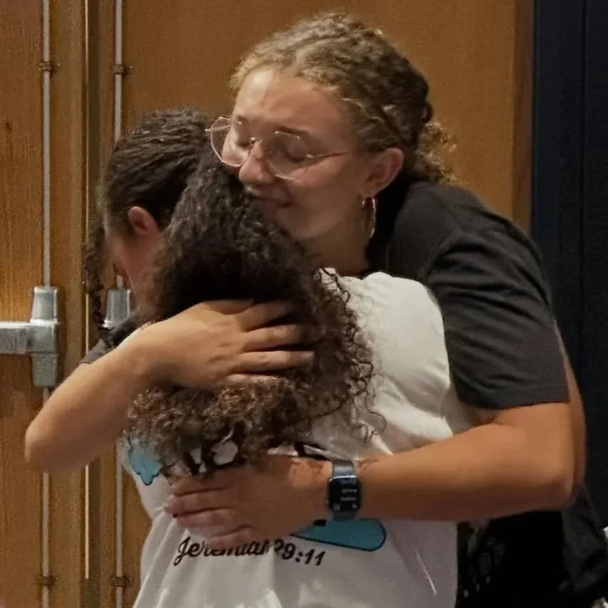 Two women hugging each other warmly, one with curly brown hair and the other with straight dark hair, in a room with wooden walls and metal pipes.