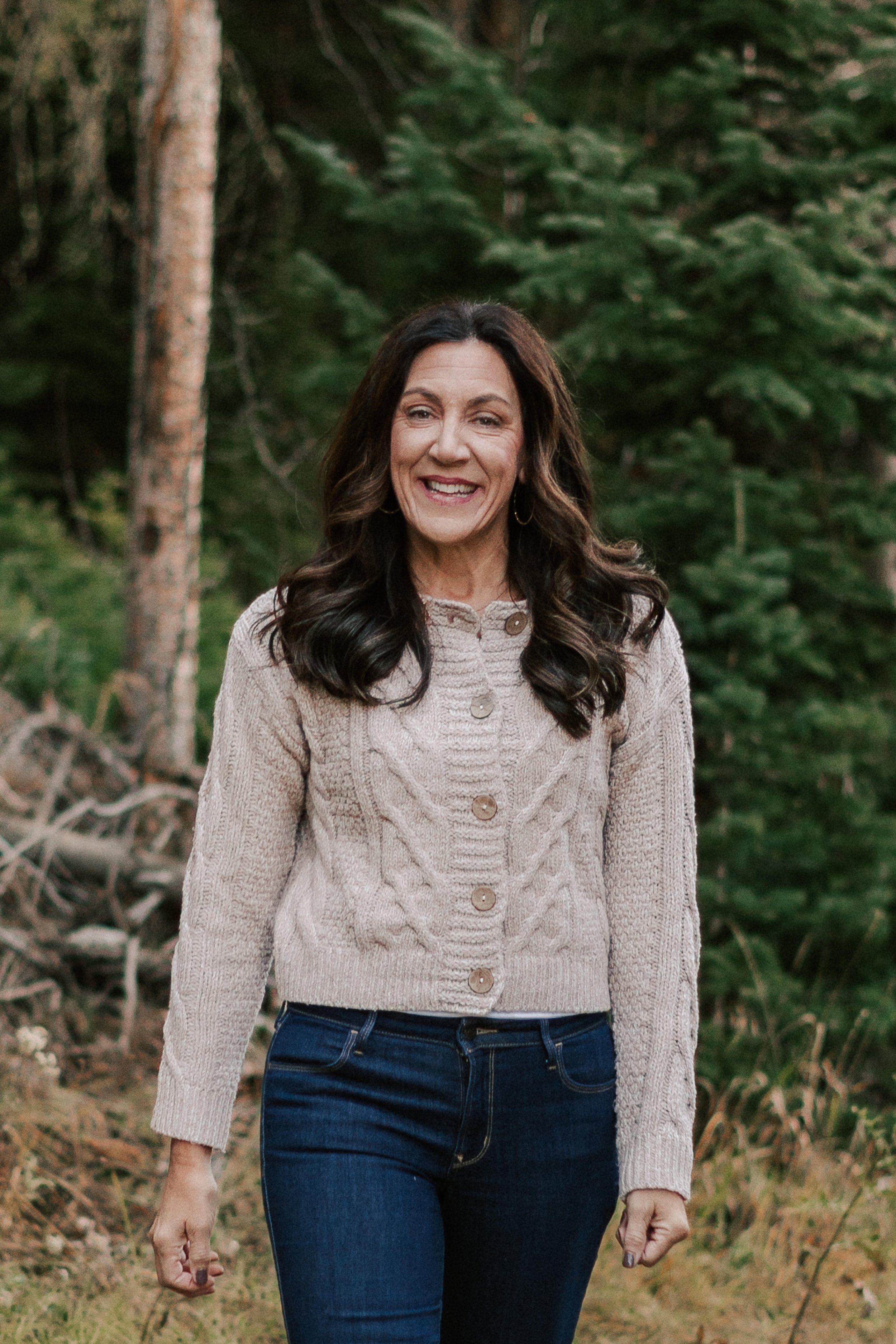 A woman smiling outdoors in a forest, wearing a beige cable-knit sweater and dark blue jeans.