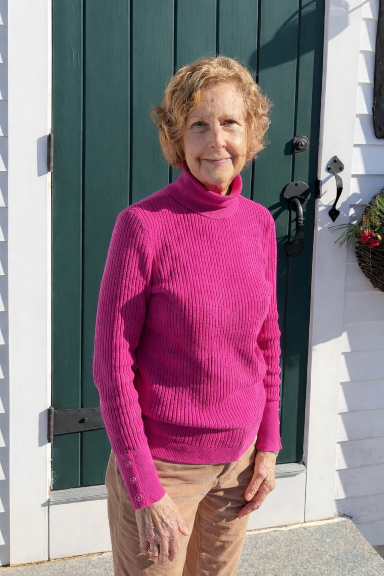 An elderly woman standing outside in front of a green door, wearing a bright pink turtleneck sweater and beige pants, smiling at the camera.