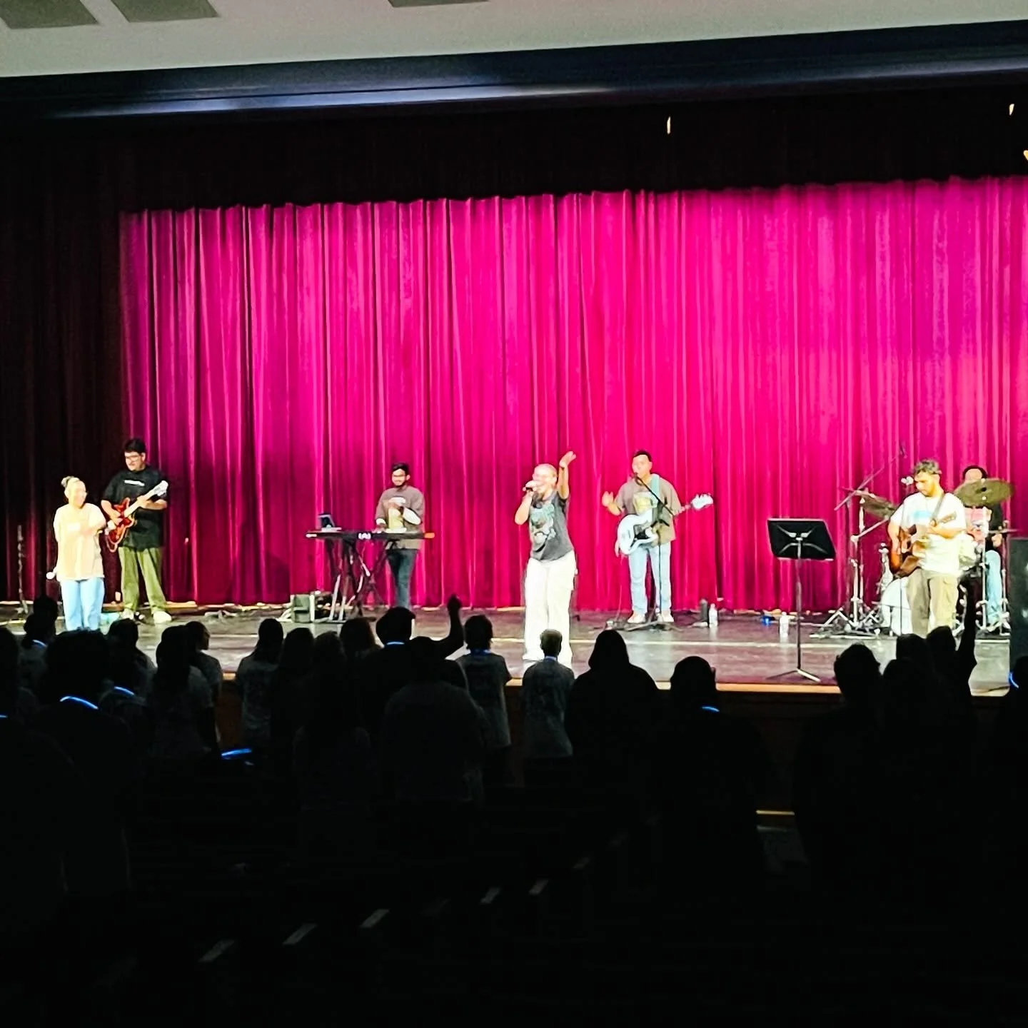 A live band performing on stage with red curtains in the background, audience standing and singing along.