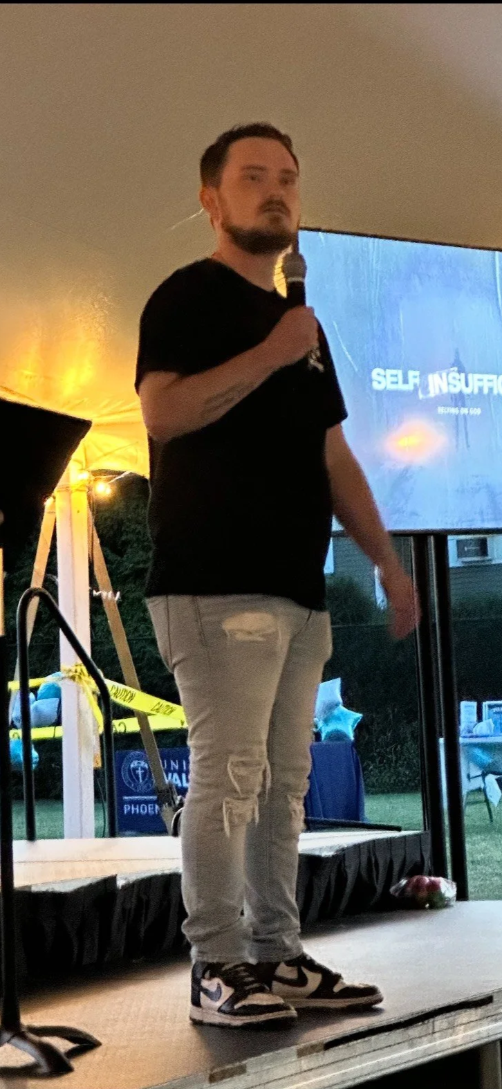Man standing on a stage speaking into a microphone at an outdoor event, with a presentation slide titled 'Self Insufficiency' projected behind him, and yellow caution tape visible in the background.